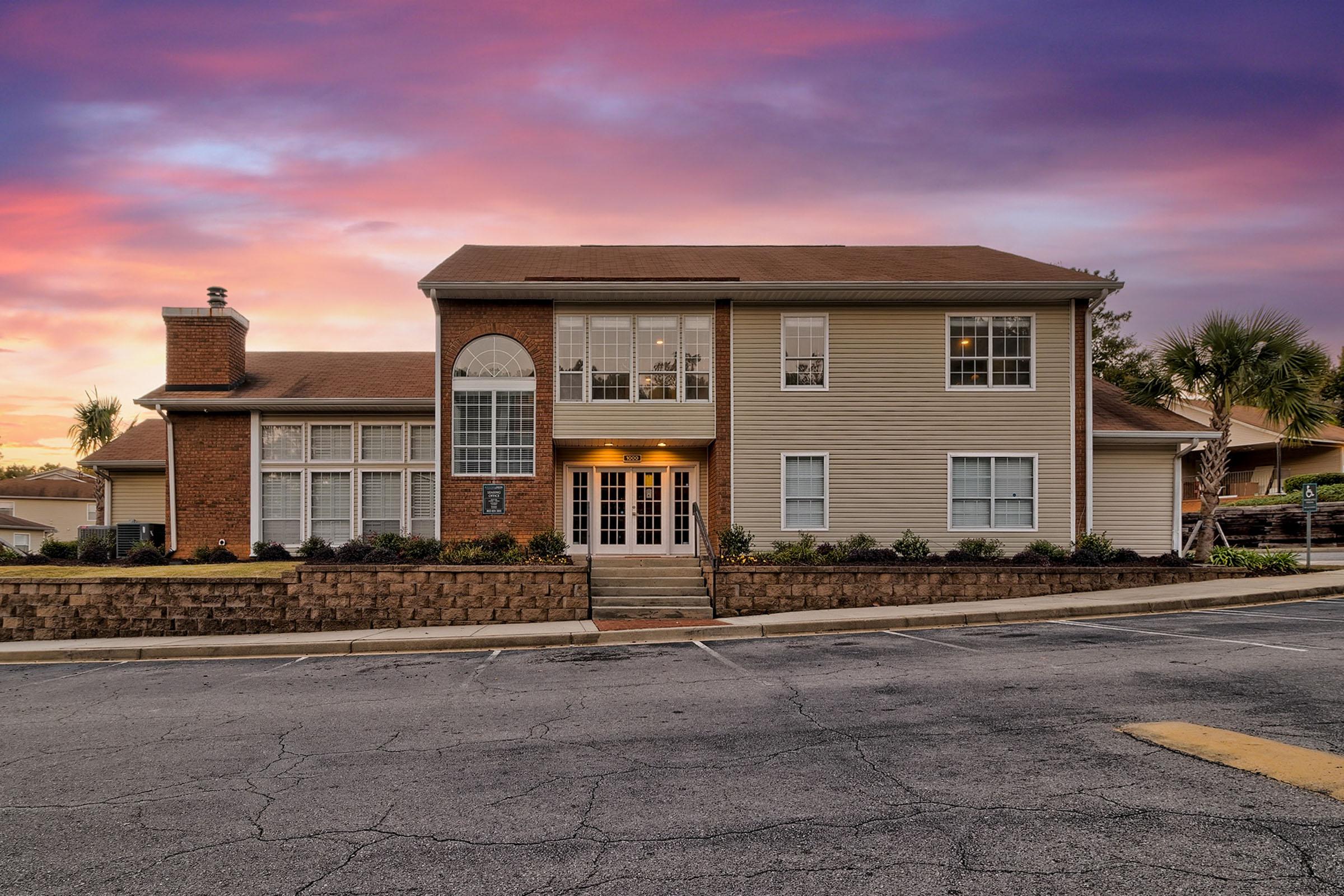 A two-story residential building with a brick and siding exterior, featuring large windows and a welcoming entrance. The sky is painted in vibrant shades of purple and orange during sunset, and the surrounding area includes landscaped greenery and palm trees.