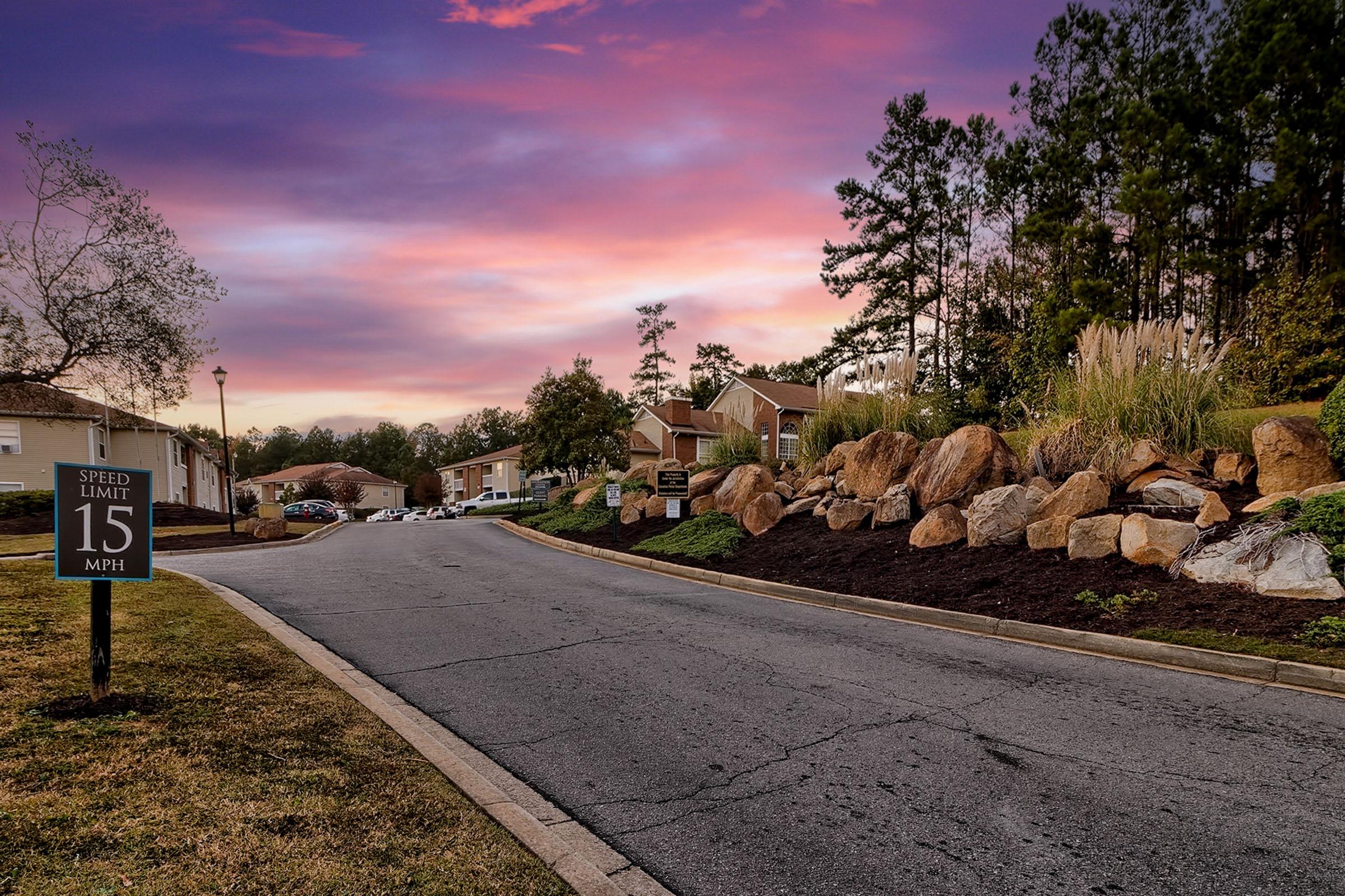 A quiet residential street at sunset, featuring a sign indicating a speed limit of 15 MPH. The road is lined with landscaped rocks and grasses, and in the background, several apartment buildings can be seen under a colorful sky with pink and purple hues.