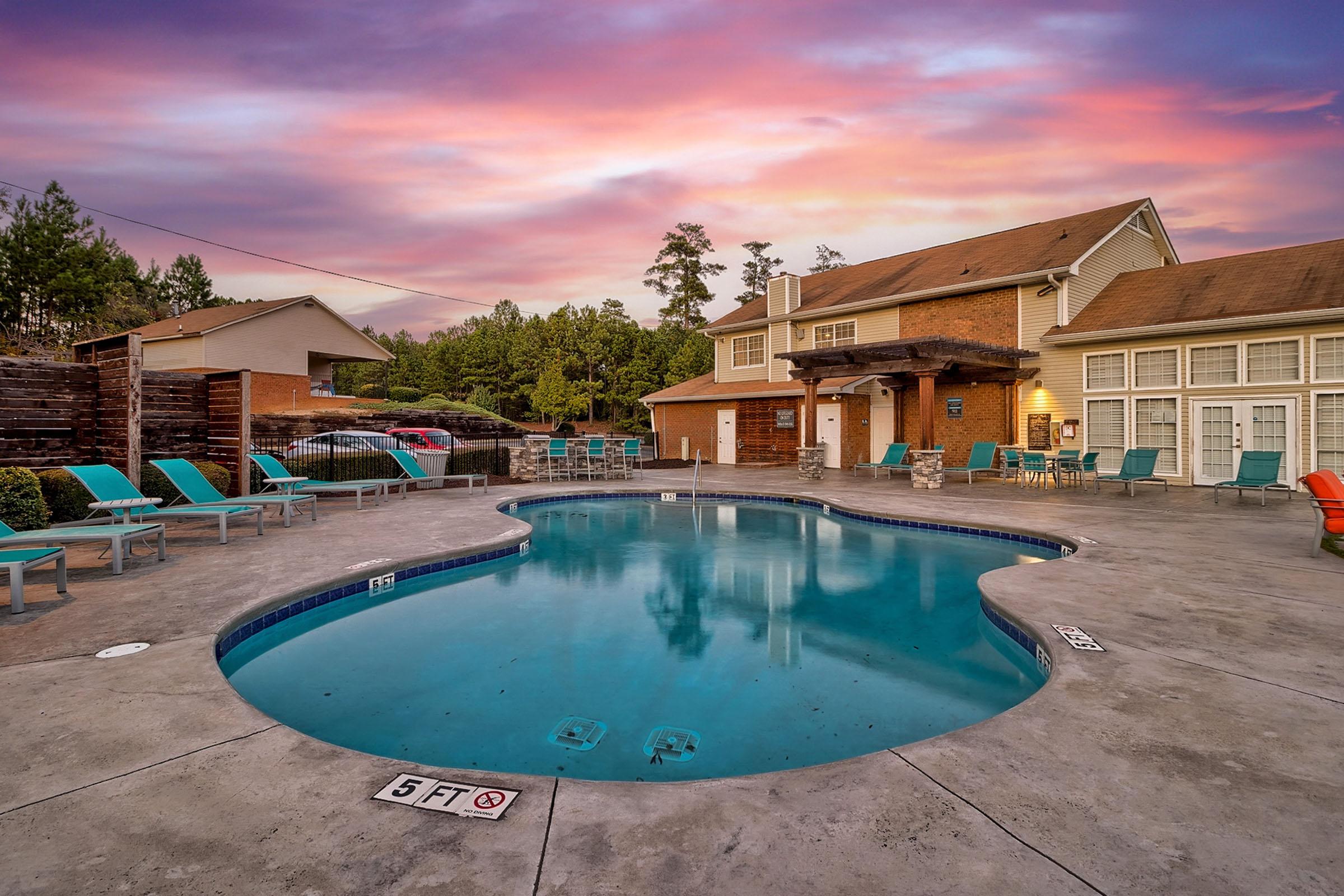 A serene outdoor pool surrounded by lounge chairs, with a vibrant sunset in the background. The area features a modern building and landscaped greenery, creating a relaxing atmosphere. The water is calm, reflecting the colorful sky, inviting guests to enjoy the tranquil setting.