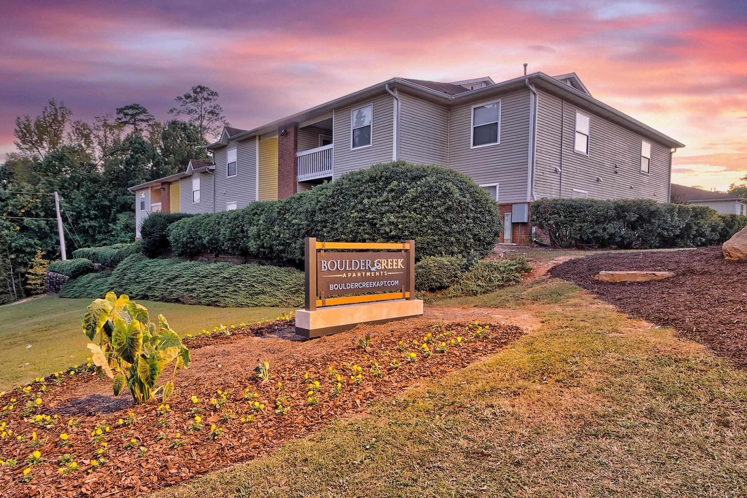 A sign for Boulder Creek Apartments is displayed in front of a well-maintained landscape featuring shrubs and flower beds. In the background, a multi-unit residential building is visible under a colorful sunset sky.