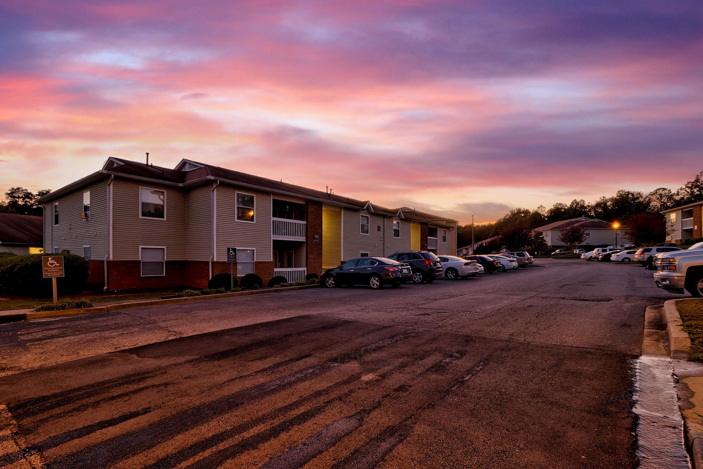 A quiet residential apartment community at sunset, with a colorful sky in shades of purple and orange. The scene features two buildings with parked cars along a paved road, highlighting a peaceful neighborhood atmosphere.