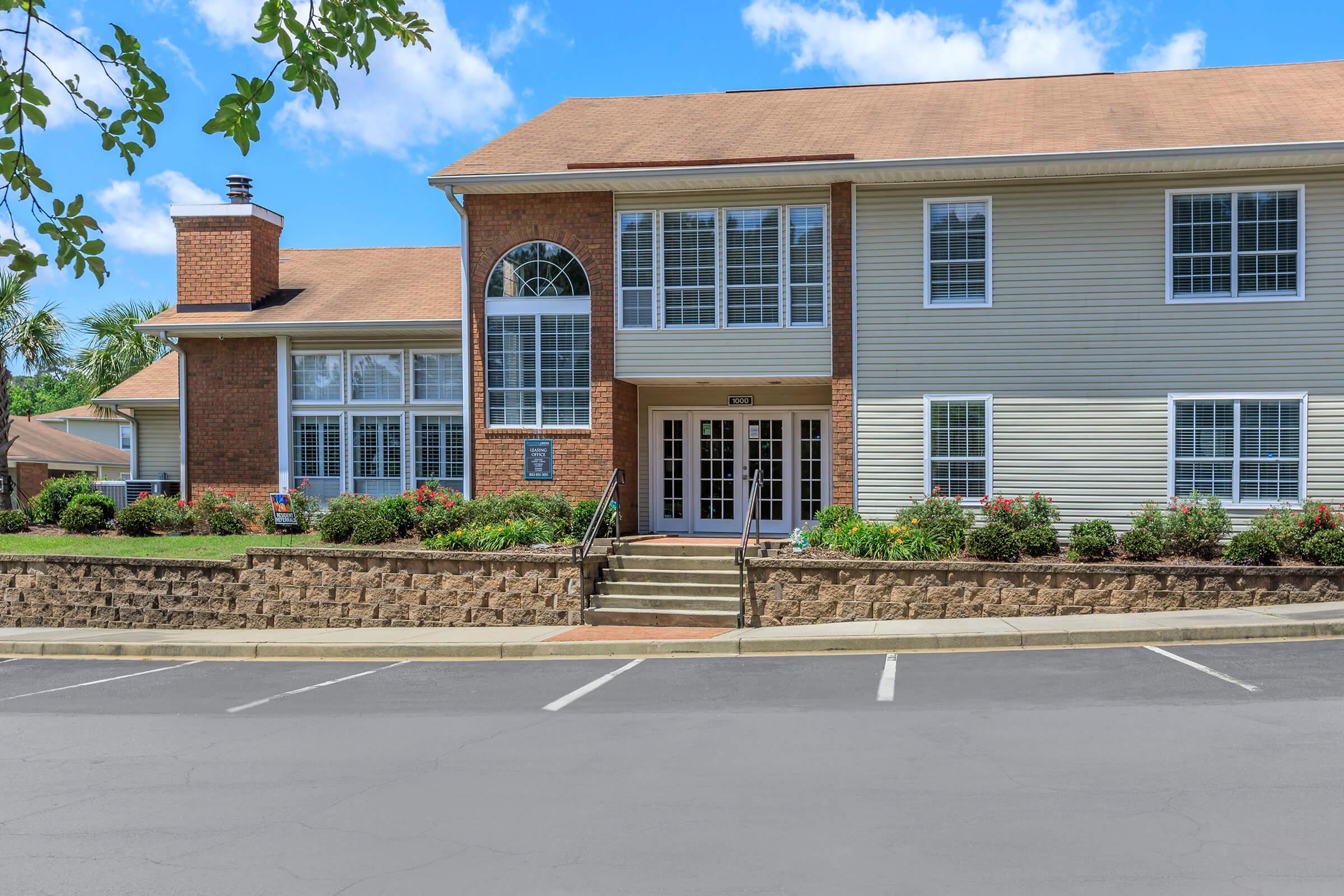 A modern, multi-unit residential building with a combination of brick and siding. The front entrance features large windows and double doors, surrounded by well-maintained landscaping and flower beds. A smooth, paved parking area is visible in the foreground, under a clear blue sky with scattered clouds.