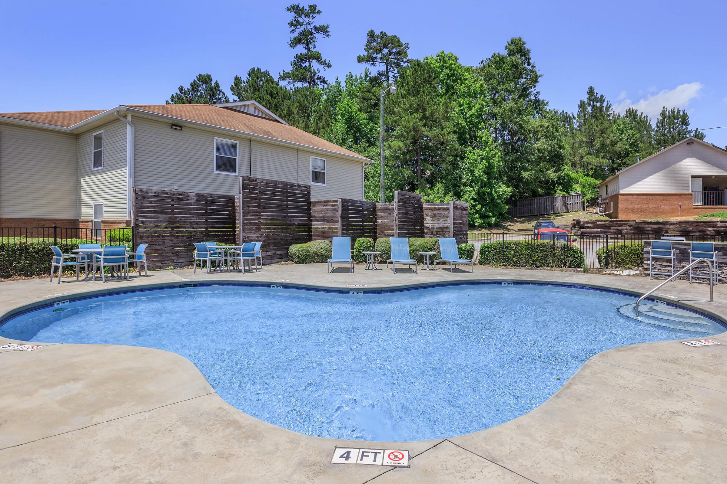 A heart-shaped swimming pool surrounded by lounge chairs and a fenced area, with a background of trees and two residential buildings. The pool has a depth marker of 4 feet visible, and the scene is set under a clear blue sky.