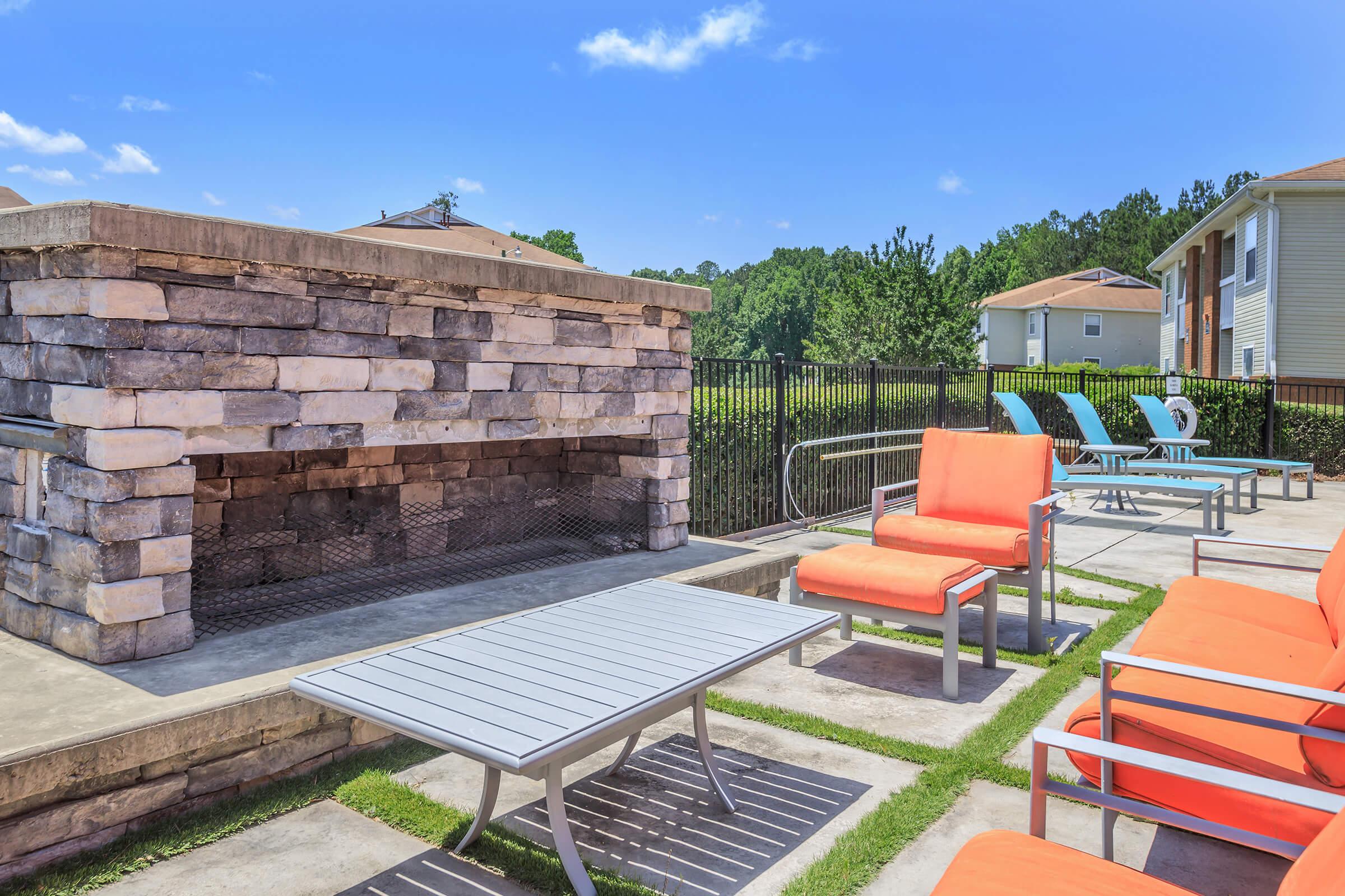 A cozy outdoor patio featuring a stone fireplace, orange cushioned chairs, and a modern table. Surrounding the fireplace are blue lounge chairs, with grass patches integrated into the design. The setting includes clear blue skies and a glimpse of residential buildings in the background.