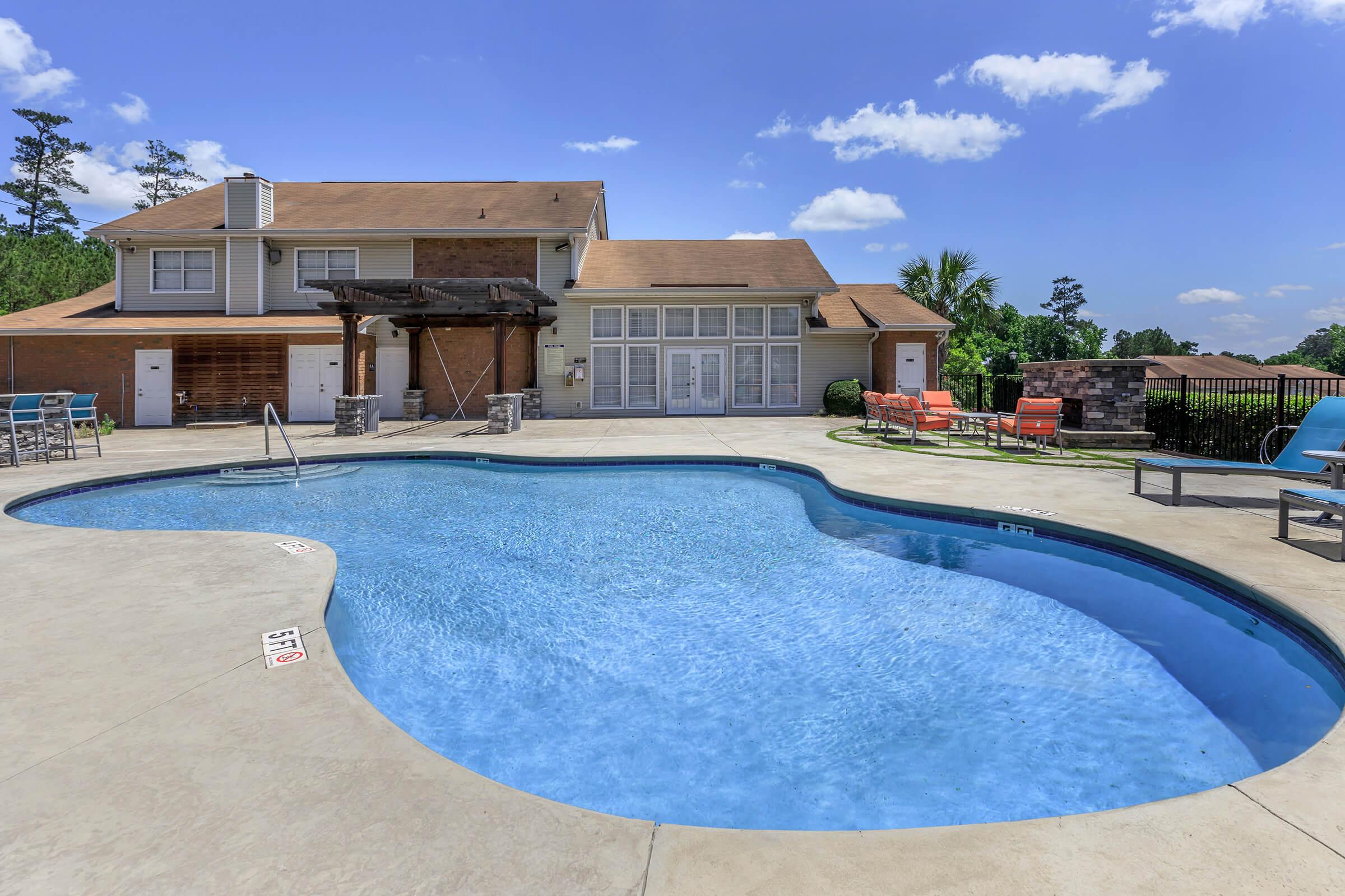 A clear, inviting swimming pool with a unique shape, surrounded by sun loungers and a patio area. In the background, there’s a two-story building with large windows, and trees in the distance. The sky is bright blue with a few clouds, creating a sunny, welcoming atmosphere.