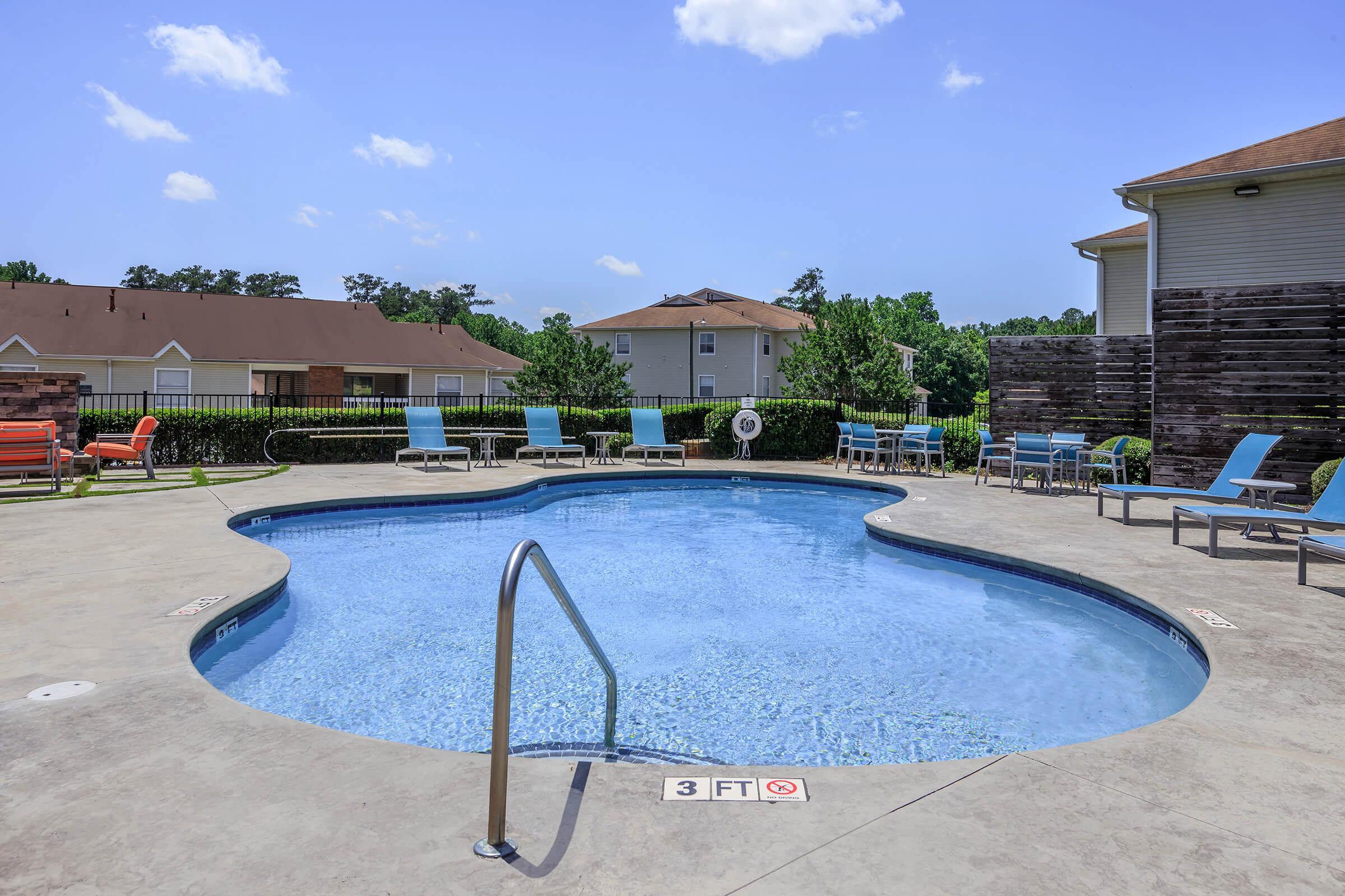 A clear swimming pool surrounded by lounge chairs and shaded areas, featuring a gradient bottom and a railing. Lush green trees and residential buildings are visible in the background under a bright blue sky with a few clouds.