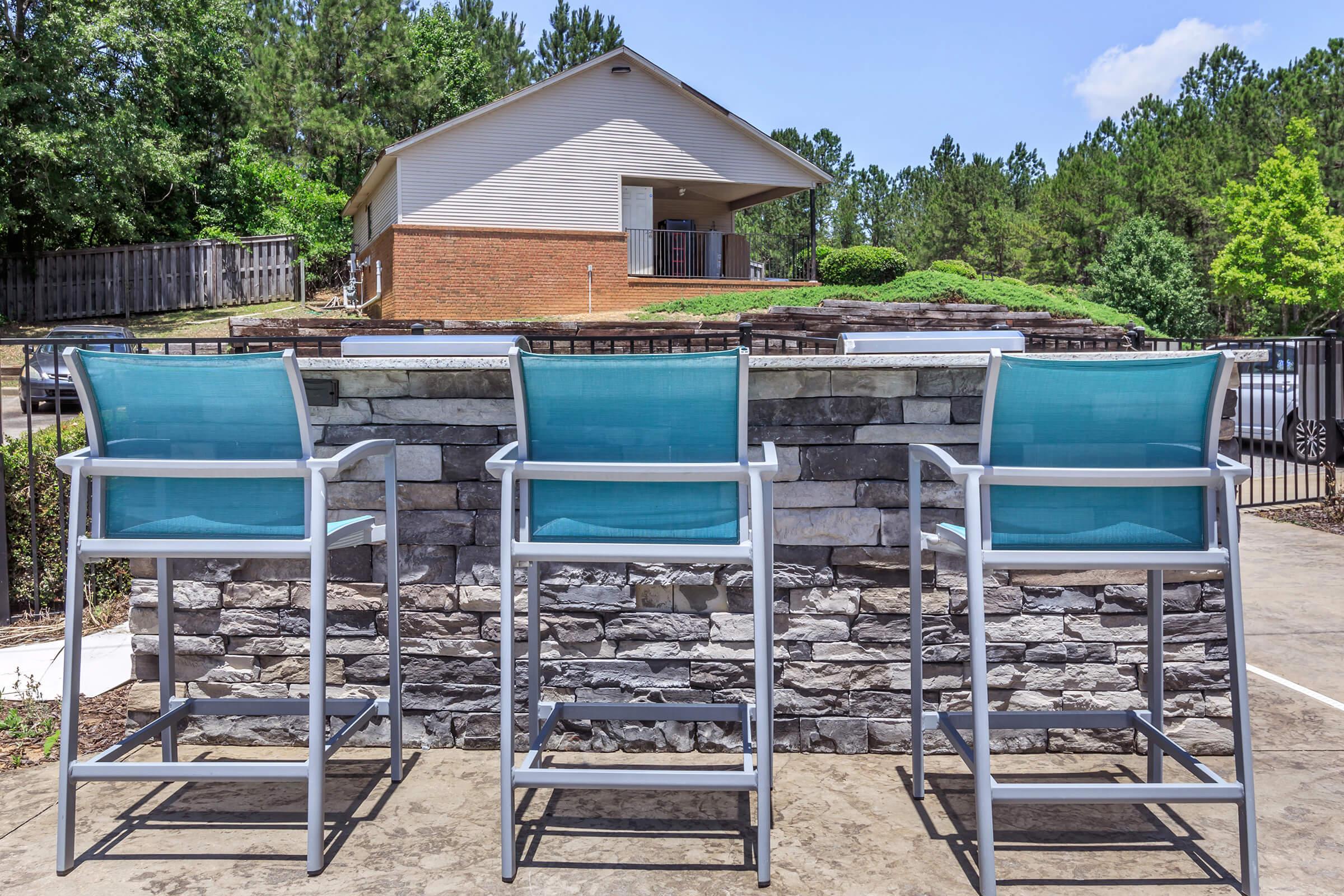 Three tall outdoor chairs with turquoise seats are lined up against a stone wall, overlooking a grassy area and a house in the background. A clear blue sky is visible above, with trees surrounding the property, creating a serene outdoor setting.