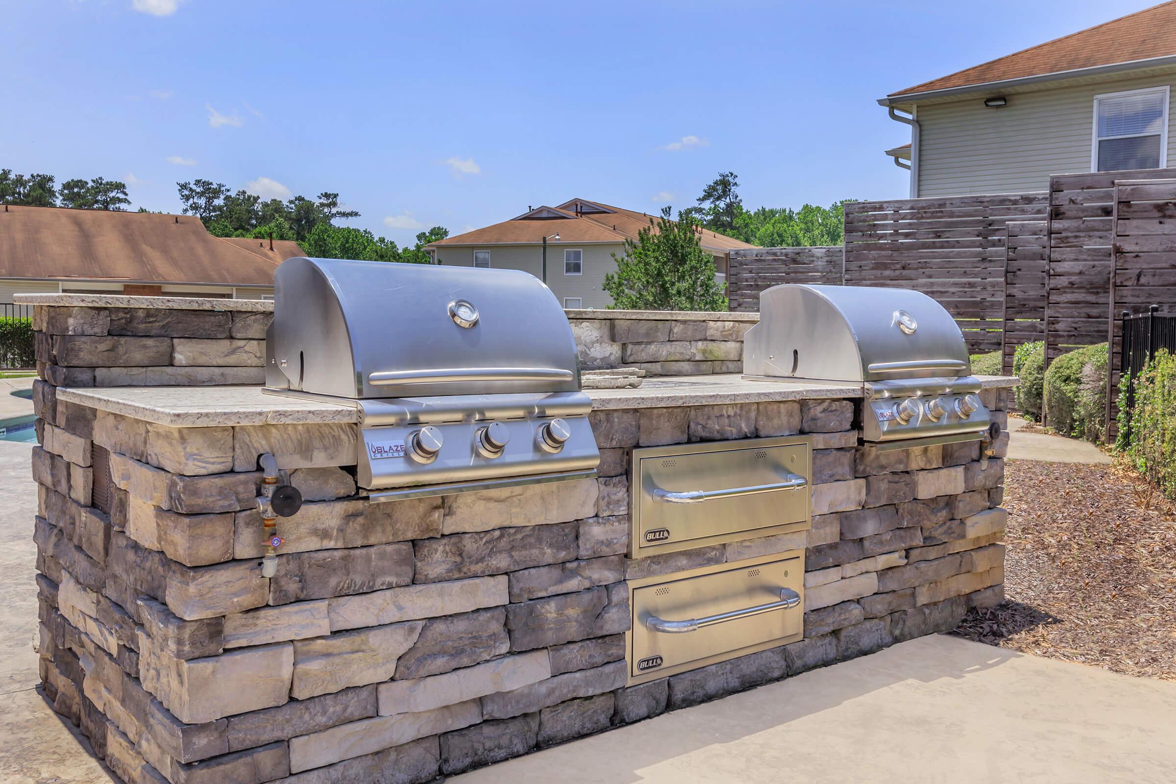 Outdoor kitchen area featuring a stone grill island with two stainless steel gas grills and a storage drawer. A landscaped yard and residential buildings are visible in the background under a clear blue sky.