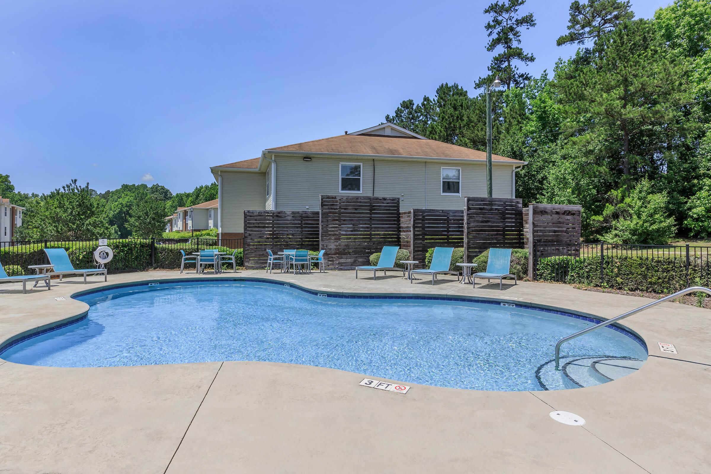 A sparkling swimming pool with a curved shape, surrounded by lounge chairs, set against a backdrop of trees and a residential building. A wooden fence provides privacy on one side, and a clear blue sky completes the serene outdoor atmosphere.