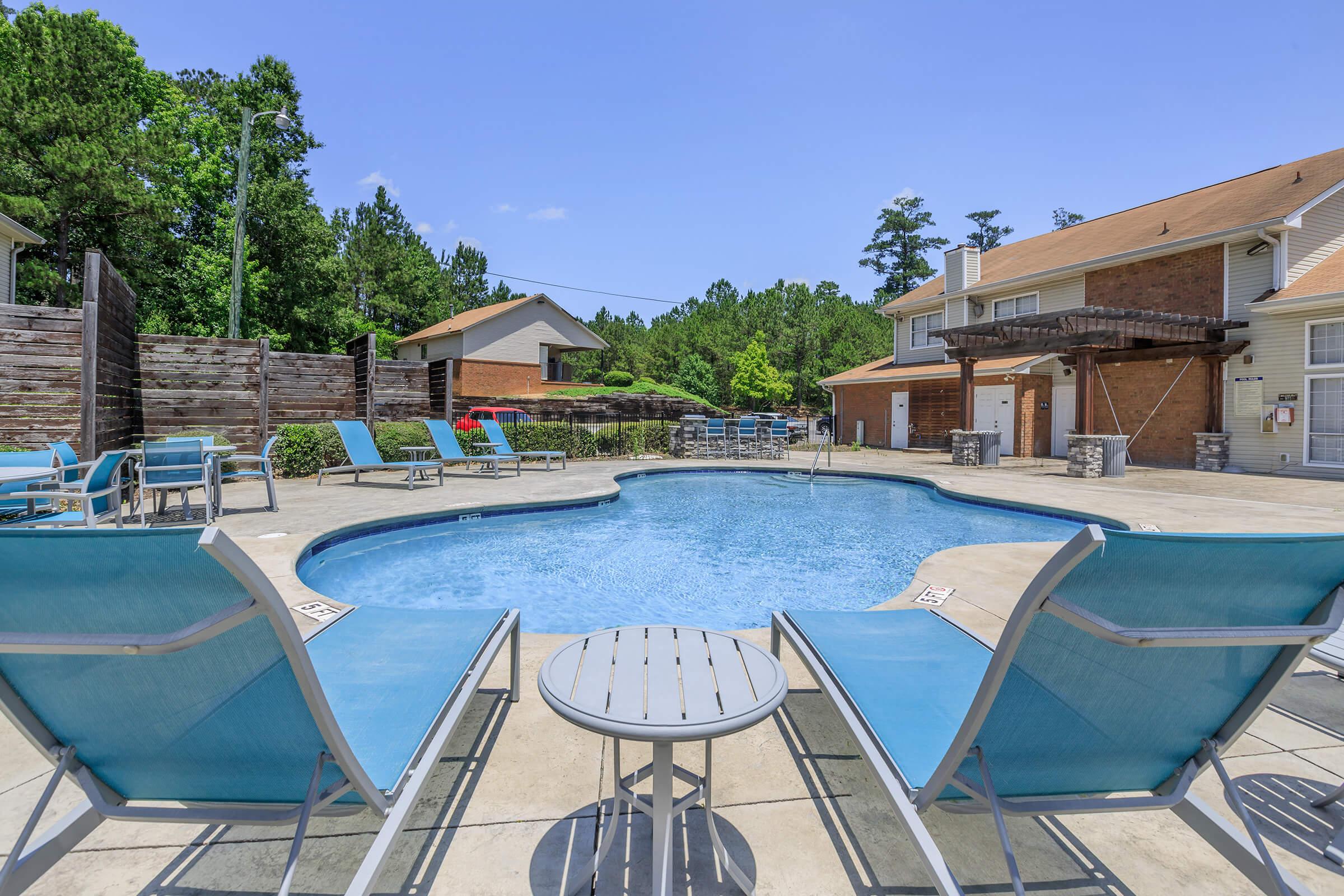 A tranquil outdoor pool area featuring a clear blue pool surrounded by lounge chairs. In the background, residential buildings and trees provide a green backdrop under a clear blue sky. Sunlight reflects off the water, creating a relaxing atmosphere for guests to enjoy.