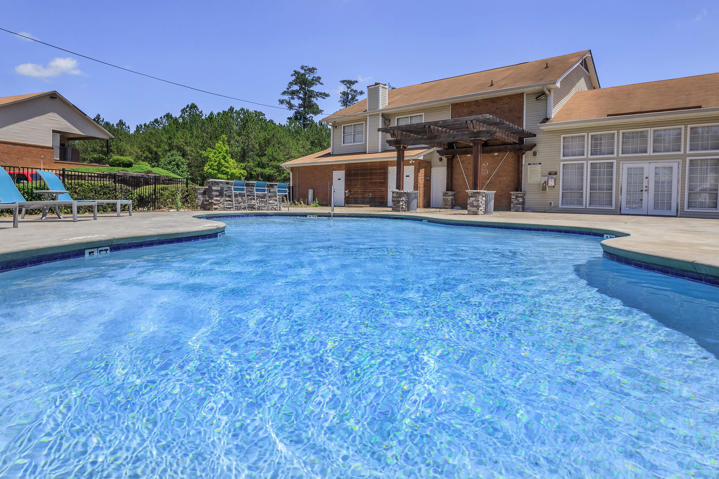 A clear blue swimming pool surrounded by lounge chairs and a wooden pergola. A two-story building with large windows is located adjacent to the pool, set against a lush, green landscape and a clear blue sky. The scene conveys a relaxing outdoor environment perfect for leisure.