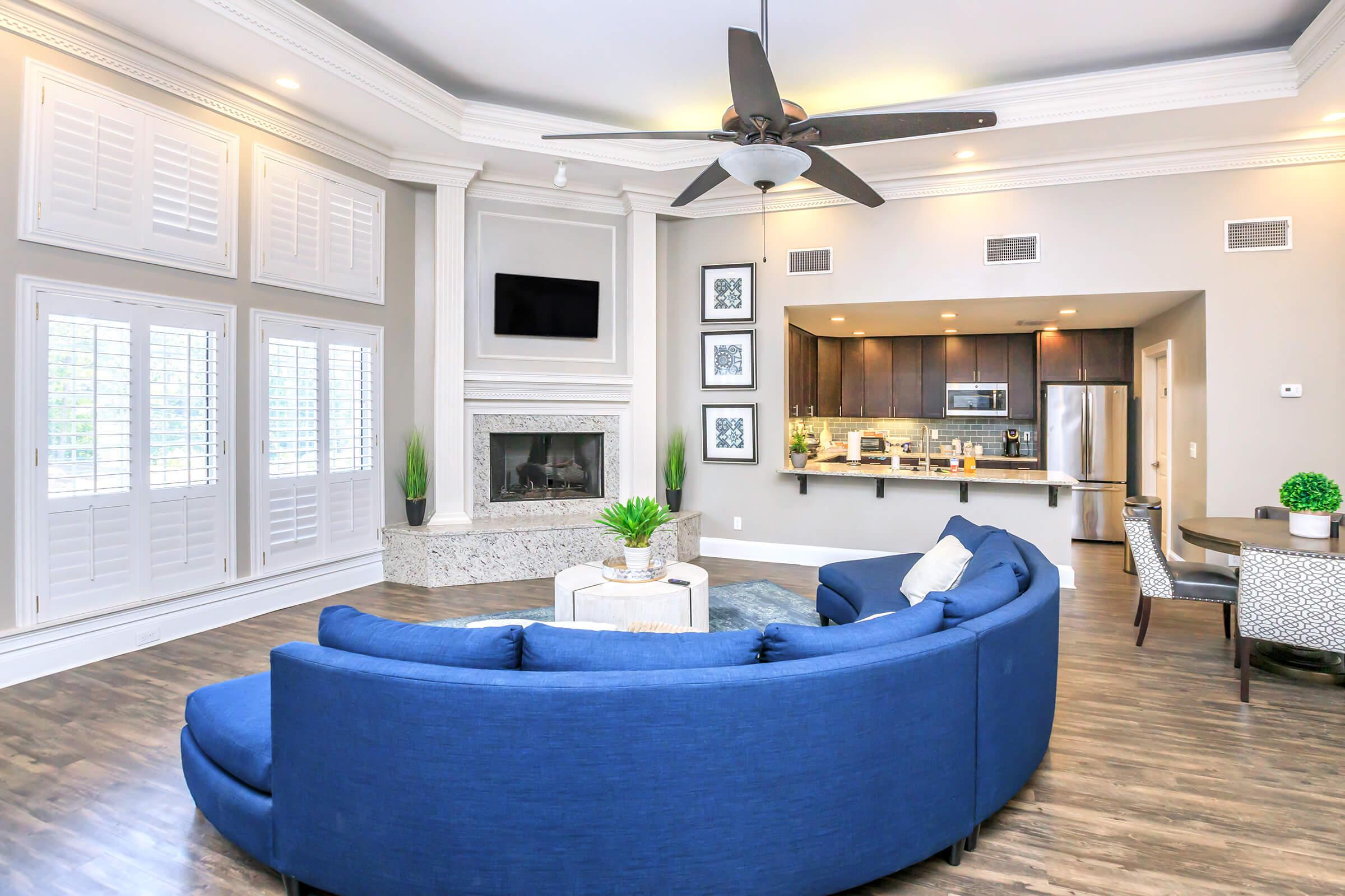 A spacious, modern living room featuring a blue curved sofa, a coffee table, and a cozy fireplace. Large windows with shutters allow natural light. The kitchen area is visible in the background, showcasing dark cabinetry and stainless-steel appliances. Elegant décor and a ceiling fan add to the contemporary ambiance.