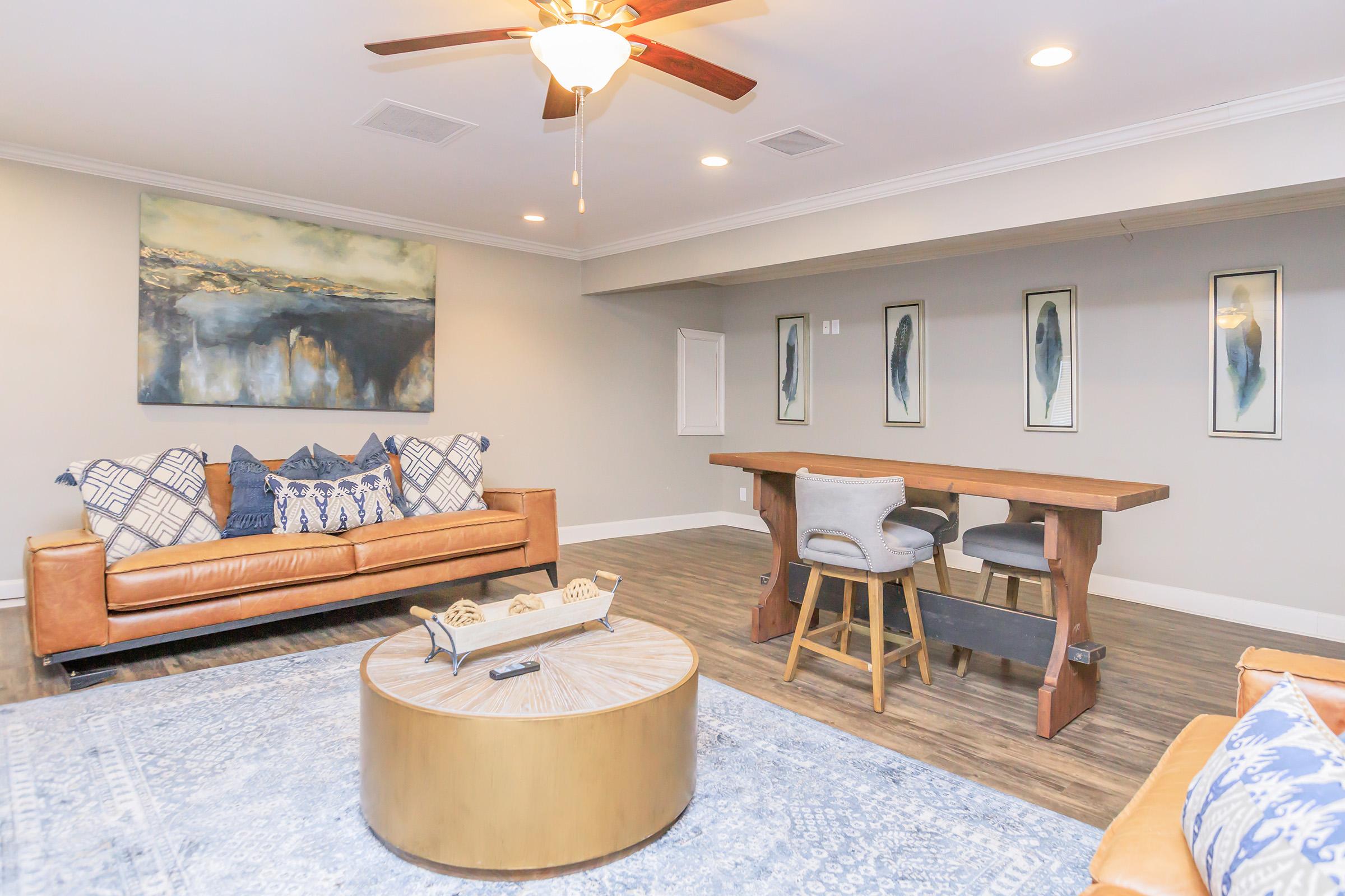 Cozy living room featuring a leather sofa with patterned pillows, a round coffee table, and a wooden bar table with stools. Decorated with abstract wall art and modern light fixtures, the space has warm lighting and a contemporary style, creating a welcoming atmosphere.