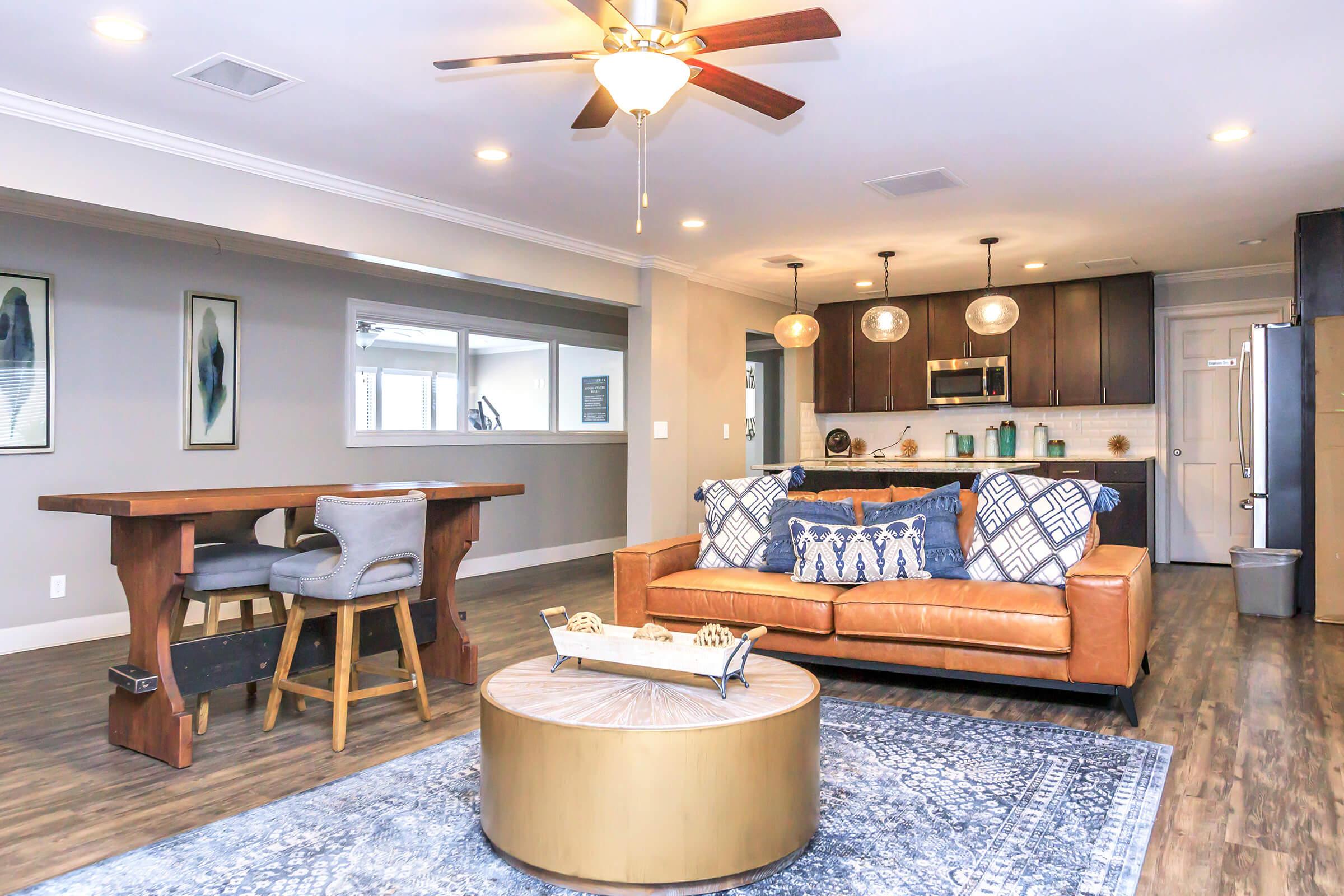 A modern living room featuring a brown leather sofa with decorative blue pillows, a round coffee table, and a wooden bar table with stools. The space has a light color scheme, wooden flooring, and stylish lighting fixtures, with a partially visible kitchen in the background.