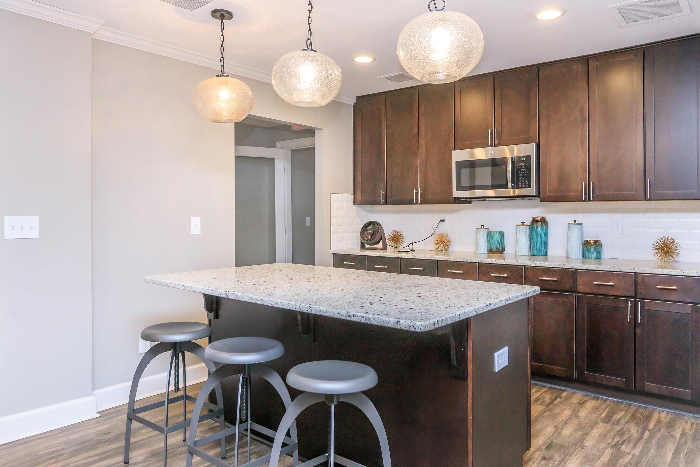 A modern kitchen featuring dark wood cabinets, stainless steel appliances, and a large countertop island with three gray bar stools. Overhead, three decorative hanging pendant lights illuminate the space. Slim blue and gold decor accents add a stylish touch to the kitchen environment.