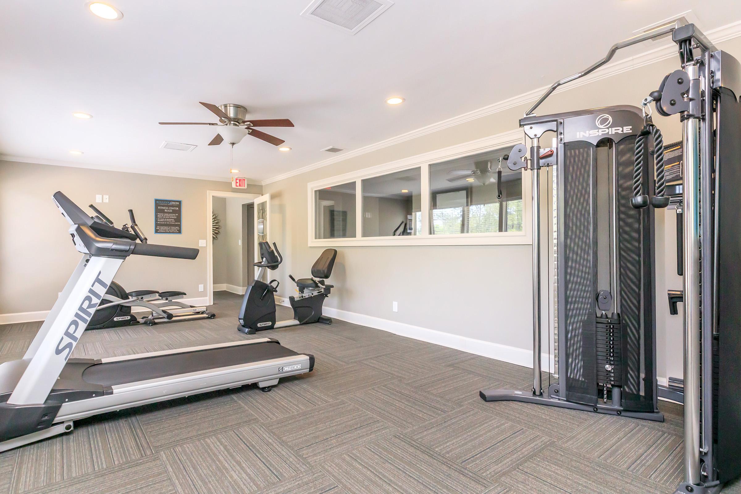 A modern gym interior featuring a treadmill, an ellipticals trainer, and a multi-station weight machine. The room is well-lit with a ceiling fan and window, showcasing a clean, organized space for workouts. The flooring is carpeted in a neutral tone, enhancing the inviting atmosphere.
