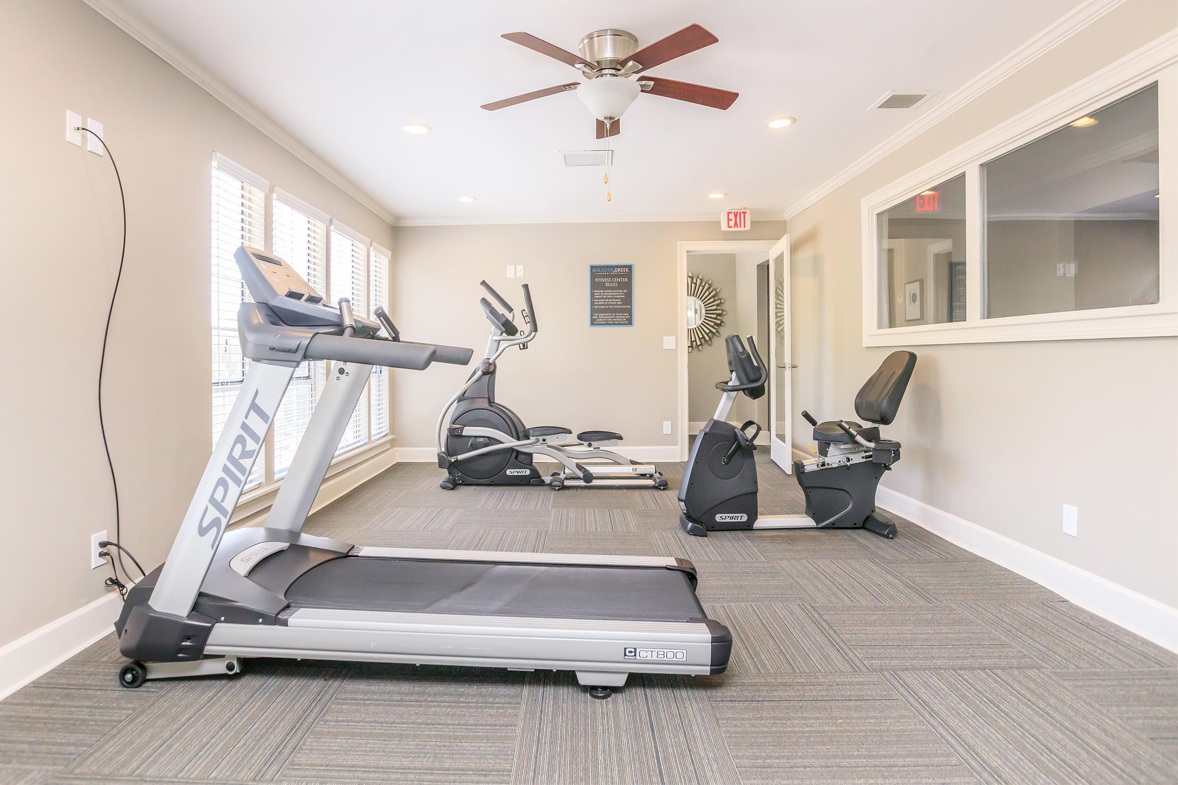 A clean, modern gym space featuring a treadmill, an elliptical machine, and a recumbent bike. The room has natural light from large windows, a ceiling fan, and neutral-colored walls, creating an inviting atmosphere for exercise.