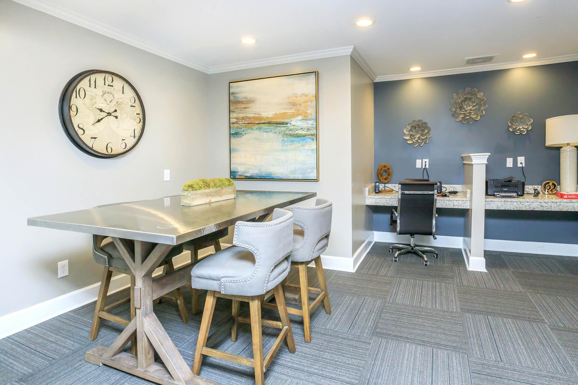 A modern office space featuring a large round clock on the wall, an abstract artwork above a sleek table with four stylish chairs. In the background, a desk with a computer is set against a dark wall, complemented by decorative wall hangings and warm lighting. The carpet is a gray, textured design.