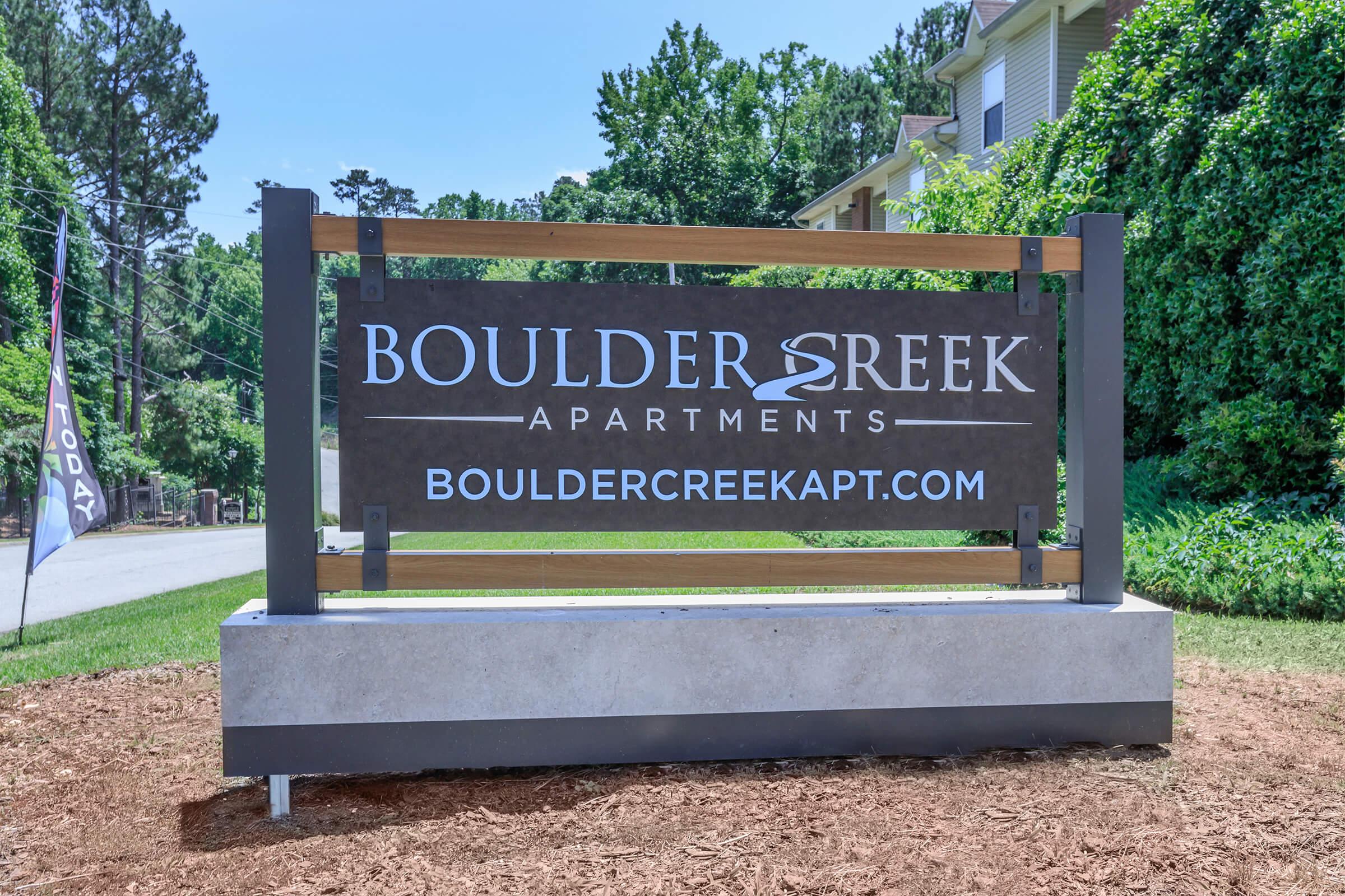 Sign for Boulder Creek Apartments displaying the name "BOULDER CREEK APARTMENTS" and the website "BOULDERCREEKAPT.COM." The sign is framed by wood and metal, with a landscaped area around it. The background features greenery and a road leading into the complex.