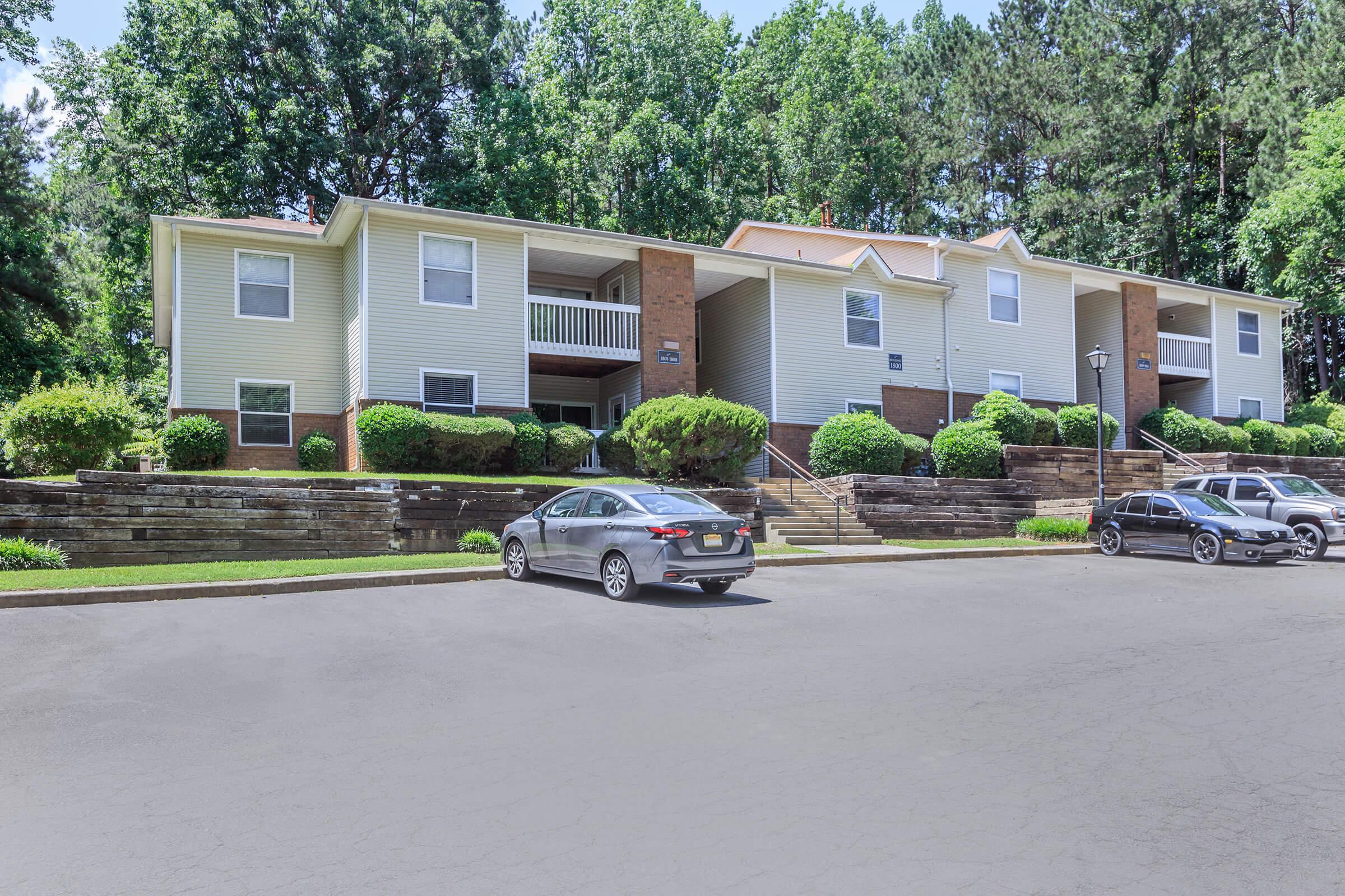 A multi-unit residential building set among green trees. The two-story structure features balconies and landscaped grounds. Several cars are parked in front of the building on a paved area, with steps leading up to the entrances. The setting is peaceful and well-maintained, showcasing a suburban environment.