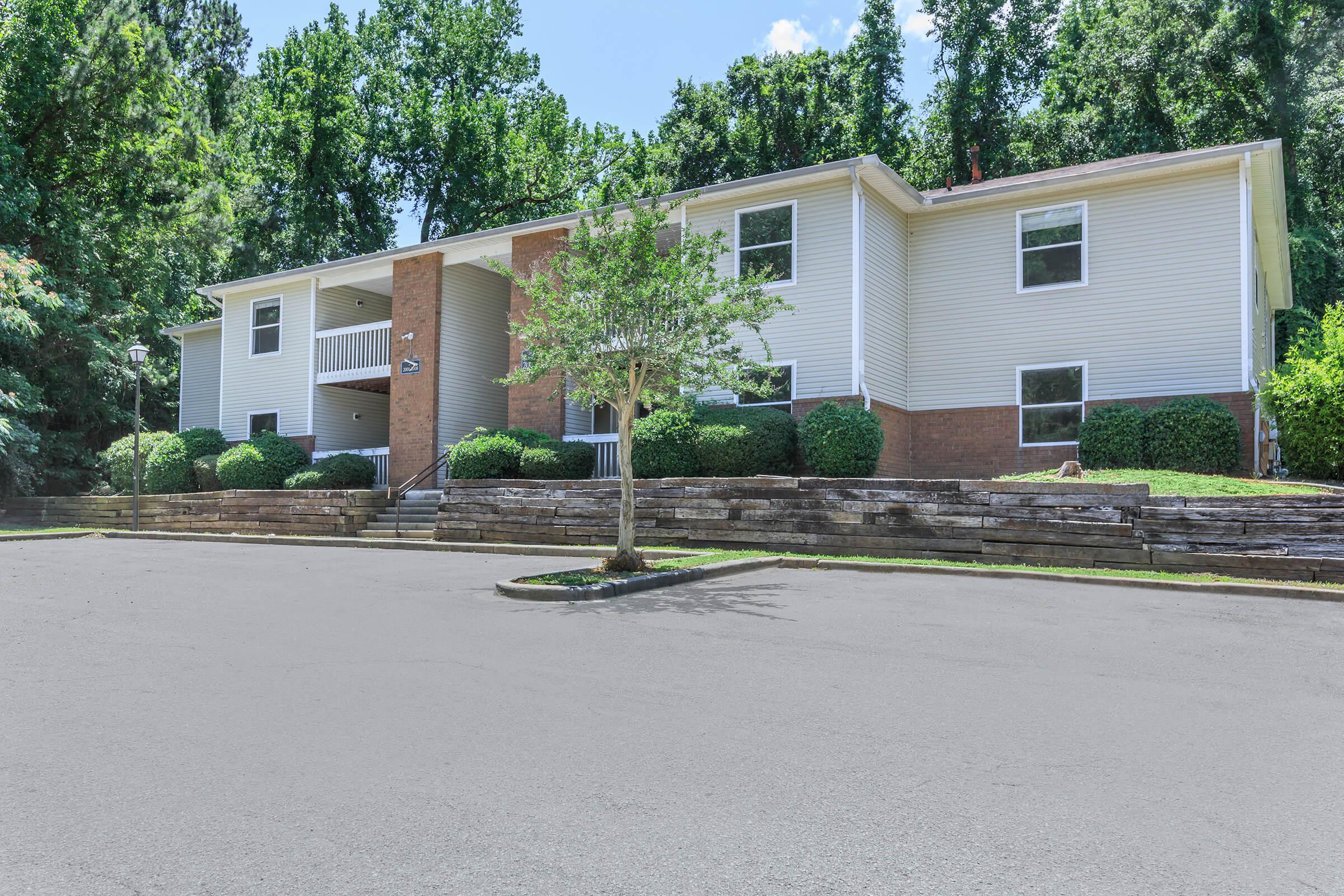 A two-story residential building with a light-colored facade, surrounded by greenery and shrubs. The entrance features a small set of stairs leading up to a covered porch. The pavement in front is clear, and trees are visible in the background, indicating a peaceful, suburban setting.