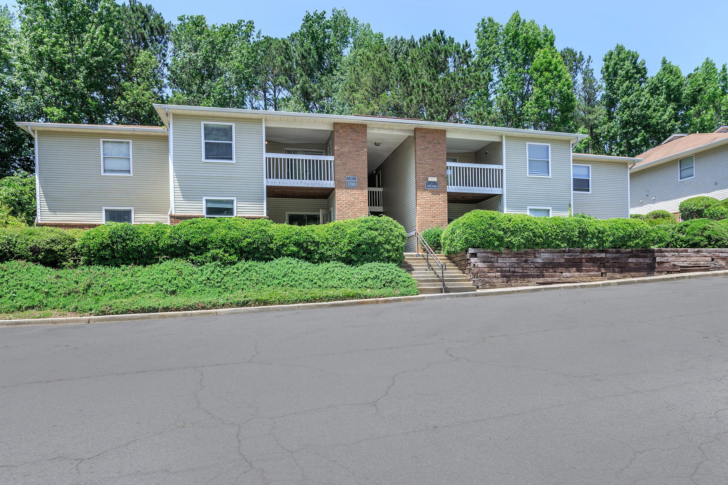 A two-story residential building with a brick entrance, surrounded by green bushes and trees. The building features balconies on both levels and is situated along a paved road. The clear blue sky adds to the bright and inviting atmosphere of the location.