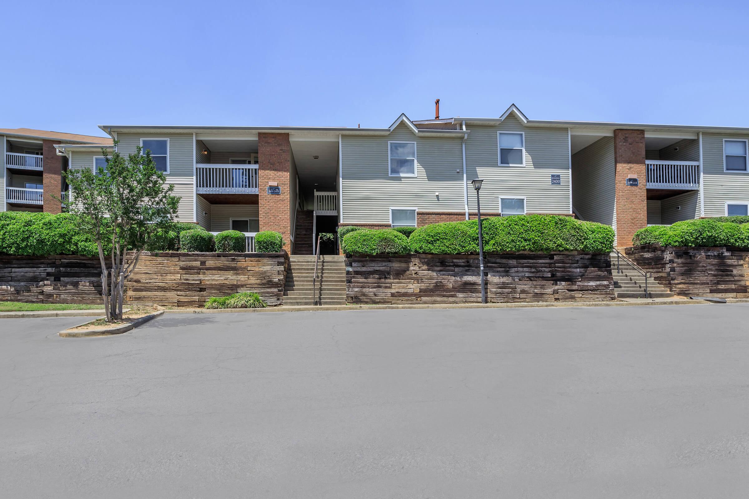 A multi-unit apartment building with two levels featuring a brick and siding exterior. The façade includes balconies and windows, surrounded by neatly trimmed bushes and landscaping. A paved road is in front of the building, with steps leading up to the entrance. The sky is clear and blue.