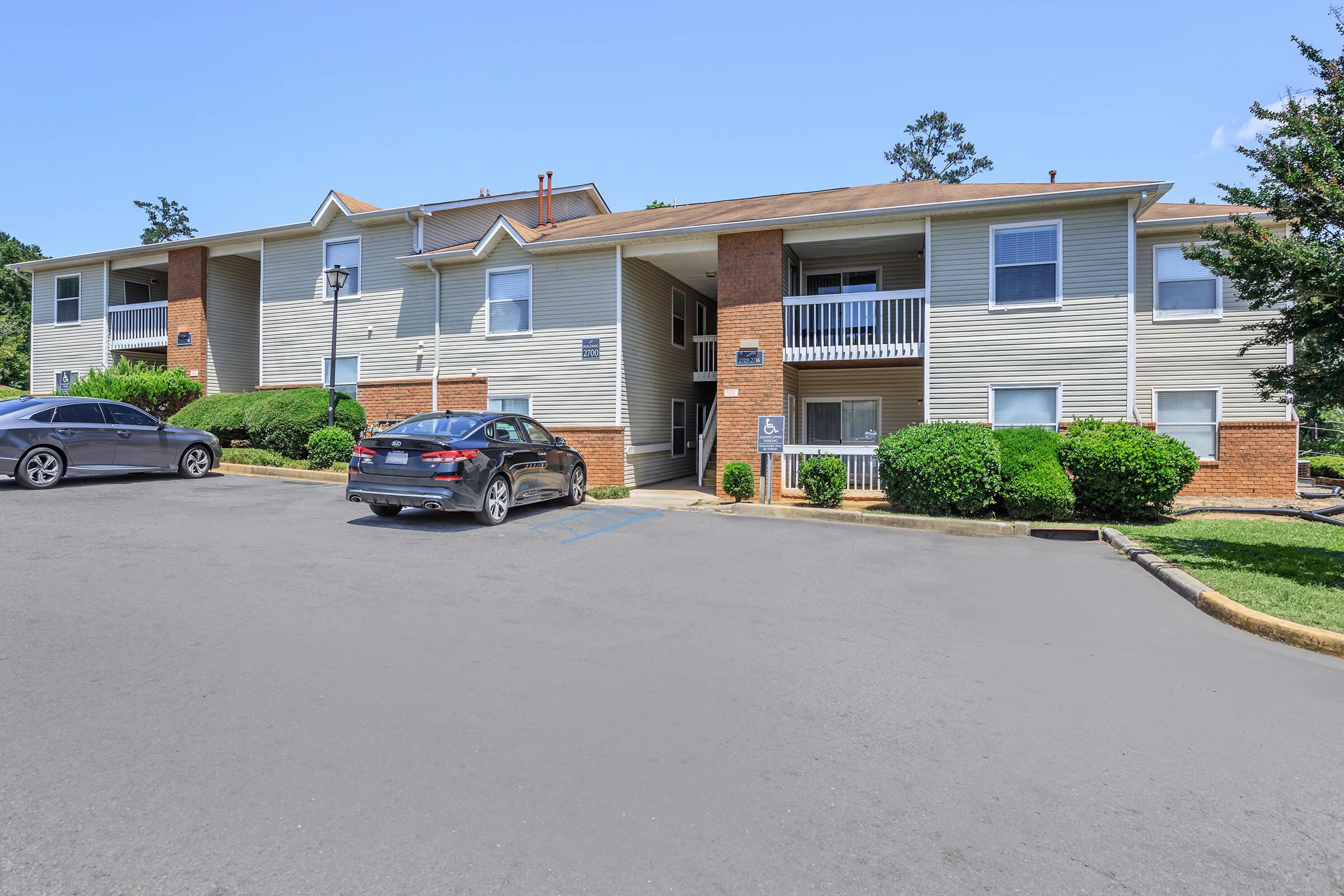 A view of a two-story apartment building with light-colored siding and brick accents, featuring balconies on the upper level. Two parked cars are visible in the foreground, with landscaped shrubs and trees surrounding the property under a clear blue sky.