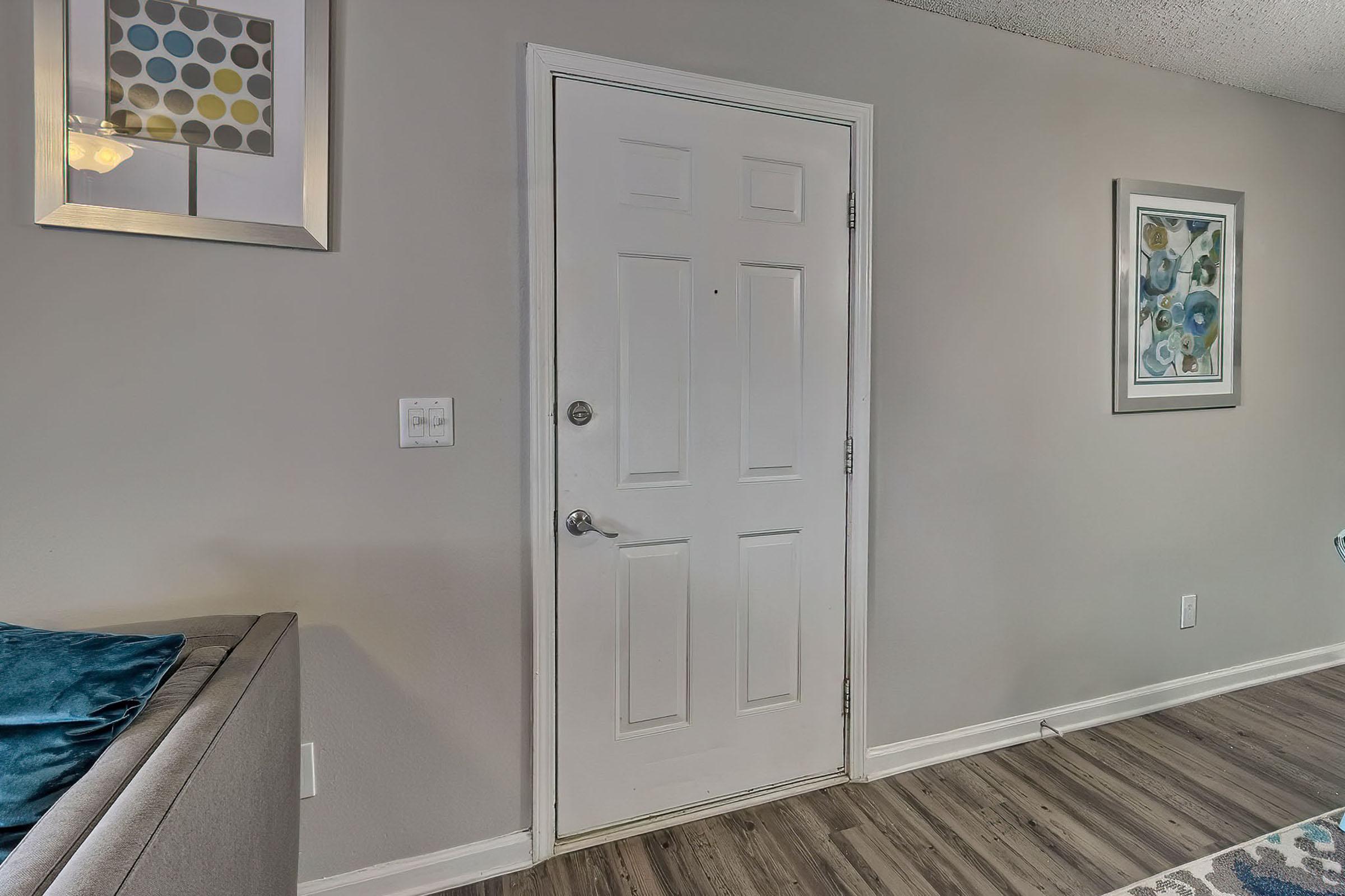 A white door with a round doorknob is located on a light gray wall. To the left, there is a gray sofa with a blue cushion. On the wall, a square framed artwork with colorful dots is displayed above a small light switch. To the right, another framed piece of artwork featuring abstract designs is visible.