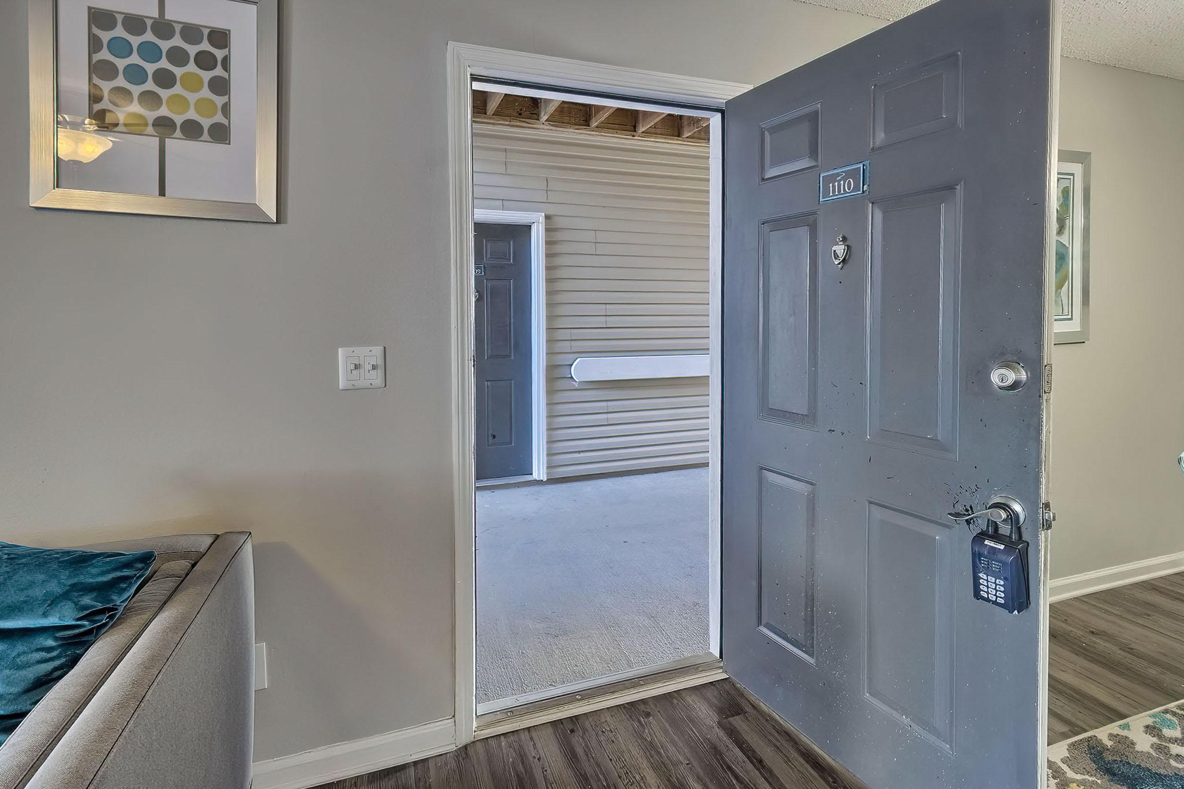 A partially open door leads to a light-filled hallway. The door has a keypad lock and a number plate. Inside, a stylish living area with a gray couch and decorative pillows is visible. The walls are painted in neutral tones, and there are framed artworks adorning the walls.