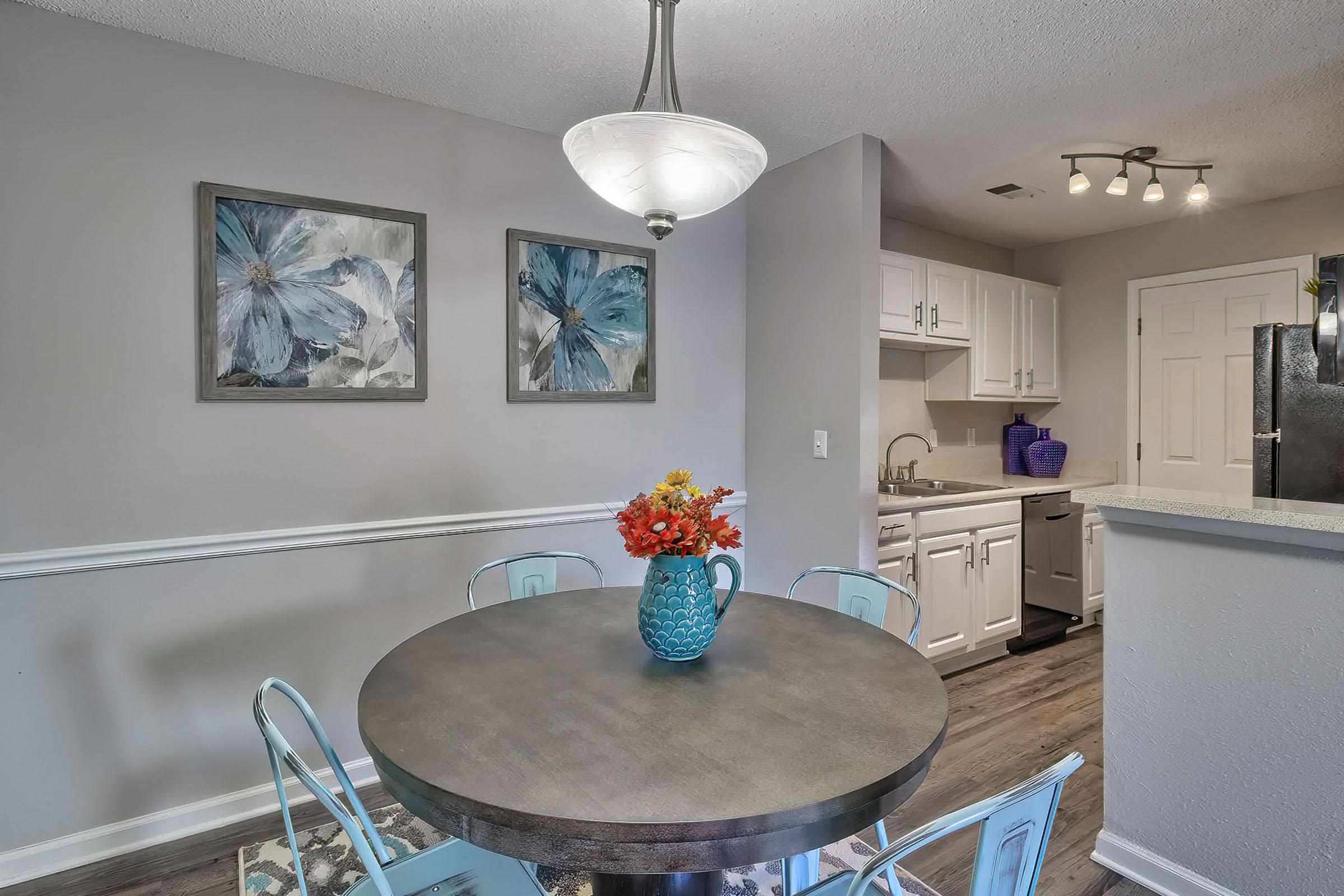 A modern kitchen and dining area featuring a round table with a blue vase of flowers, blue chairs, and two floral artworks on the wall. The kitchen includes white cabinets, a stainless steel refrigerator, and a light fixture above the table. The overall decor is bright and inviting.