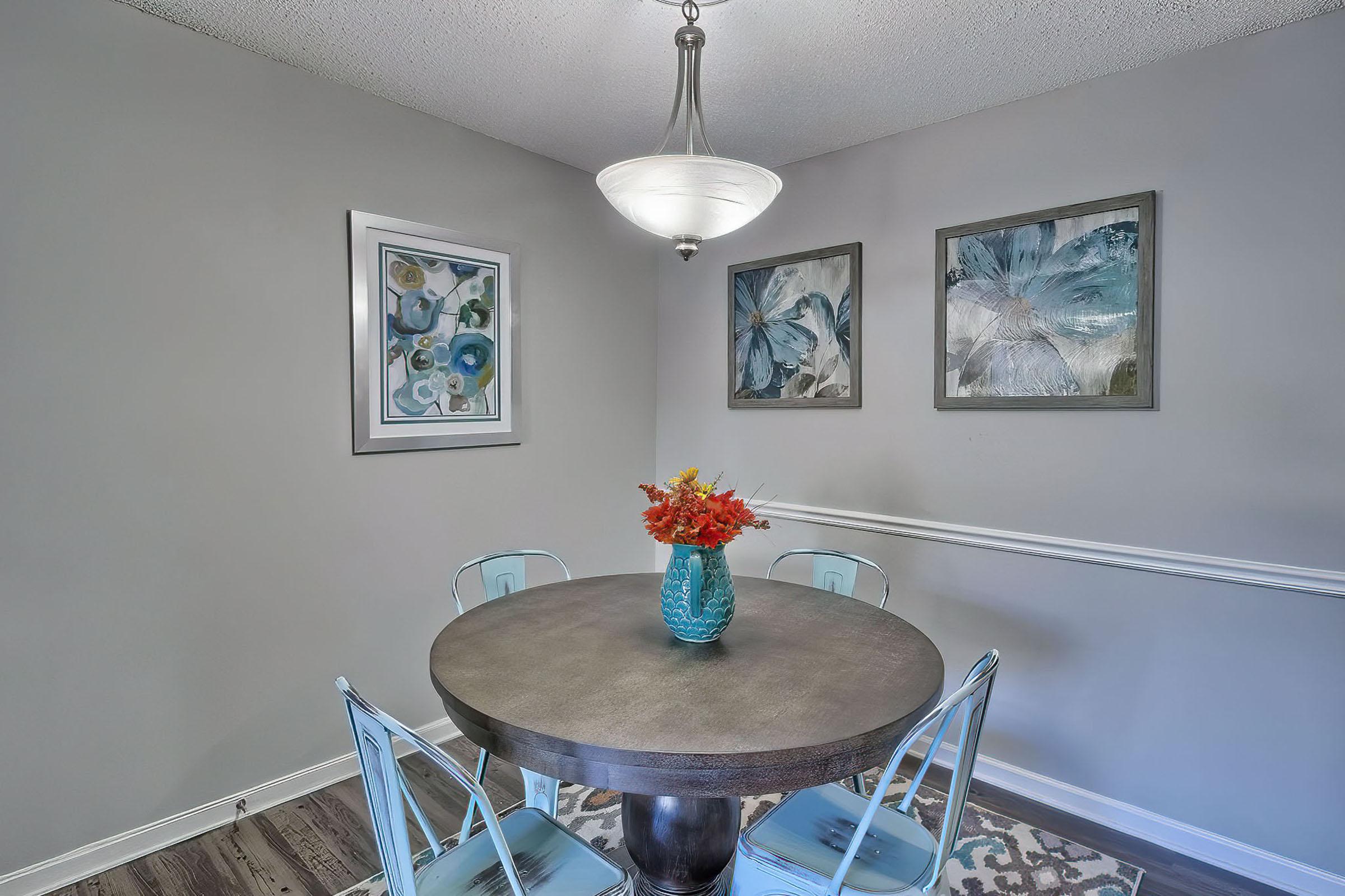 A modern dining area featuring a round wooden table surrounded by four light blue chairs. A turquoise vase filled with colorful flowers sits at the center of the table. The walls are painted gray, and two pieces of abstract art are framed and hung on the wall, alongside a pendant light hanging from the ceiling.