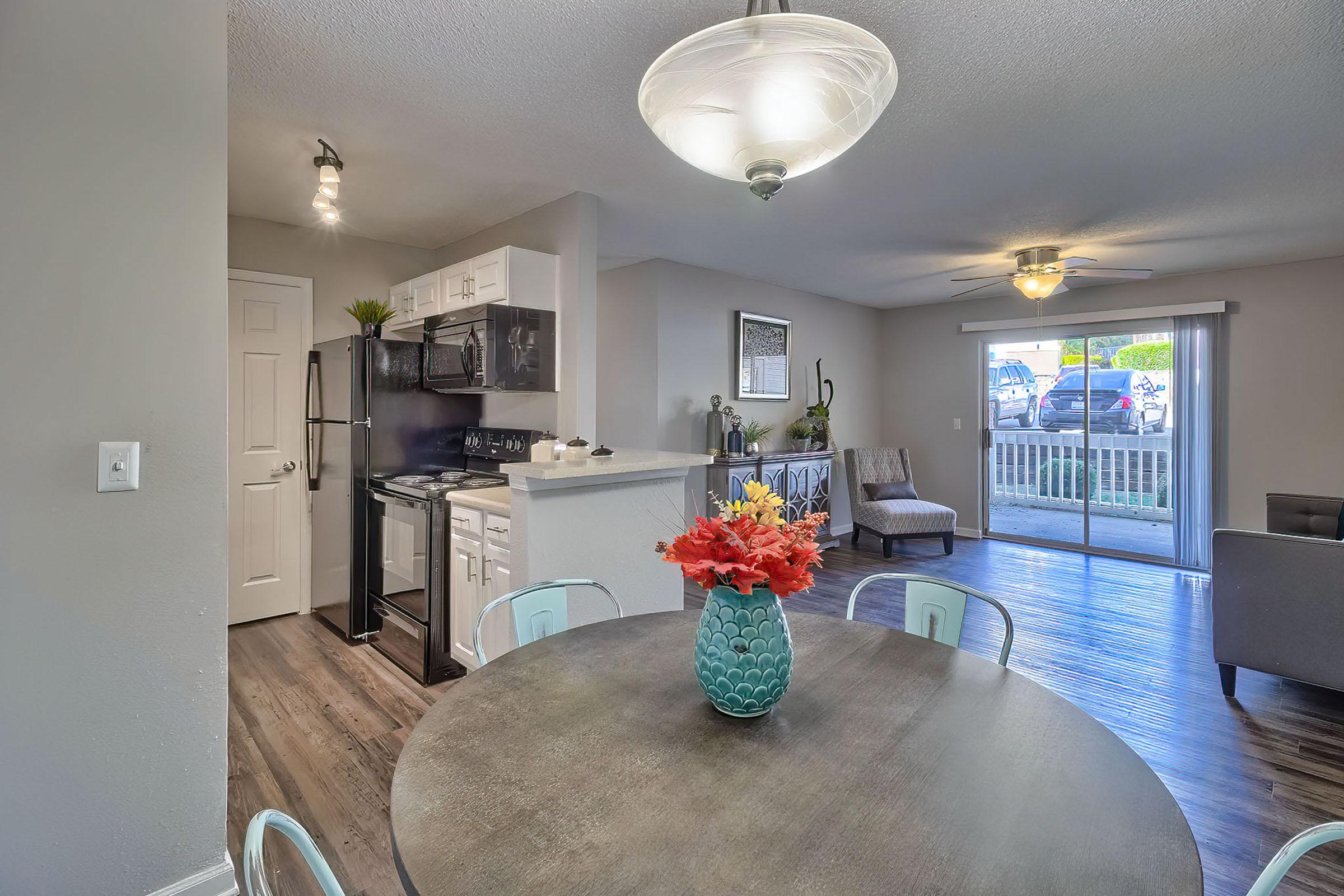 A modern living space featuring a round dining table with a vase of flowers, a kitchen with black appliances and white cabinetry, and a cozy sitting area with a gray couch. Large windows allow natural light to fill the room, and there is a ceiling fan in the living area, creating a welcoming atmosphere.