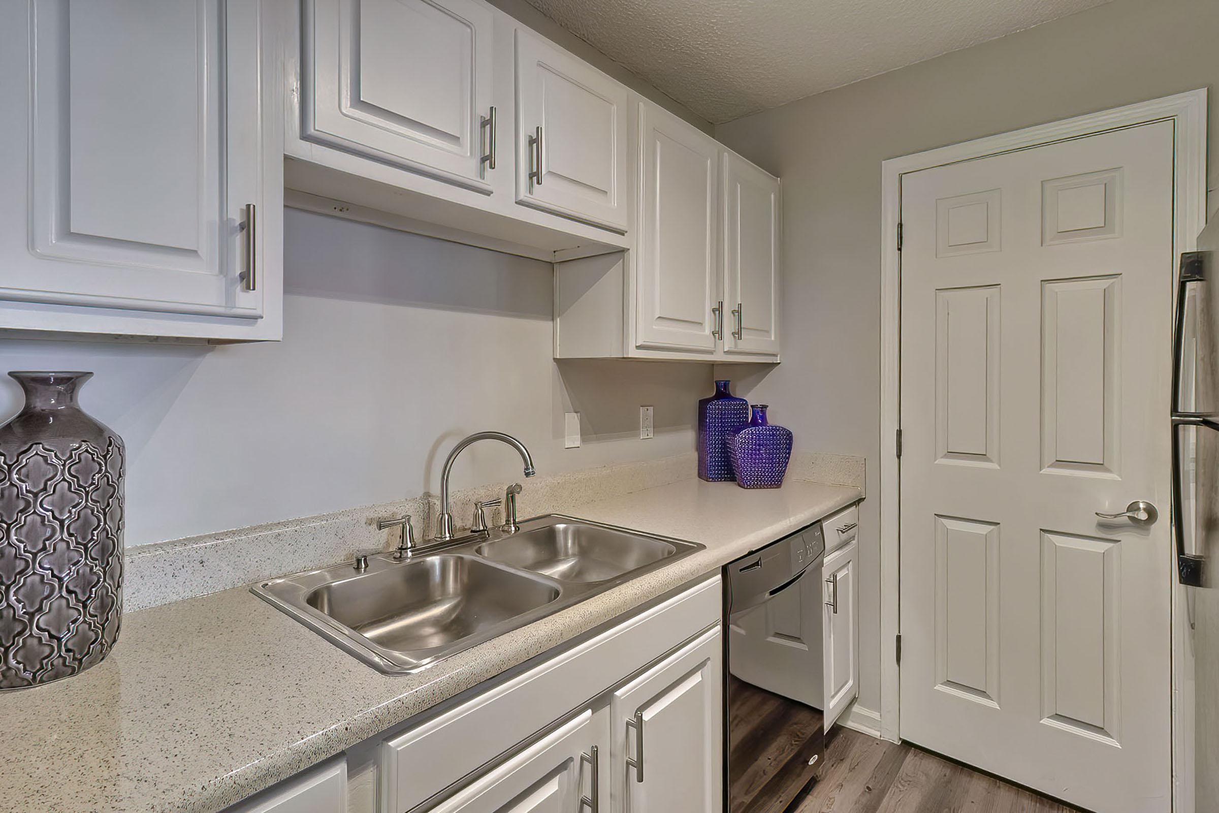 A modern kitchen featuring white cabinets, a light countertop, and a stainless steel sink with a polished faucet. Two decorative purple vases are placed on the counter, and a closed door leads to another area. The flooring has a warm, wooden appearance, contributing to the contemporary design.