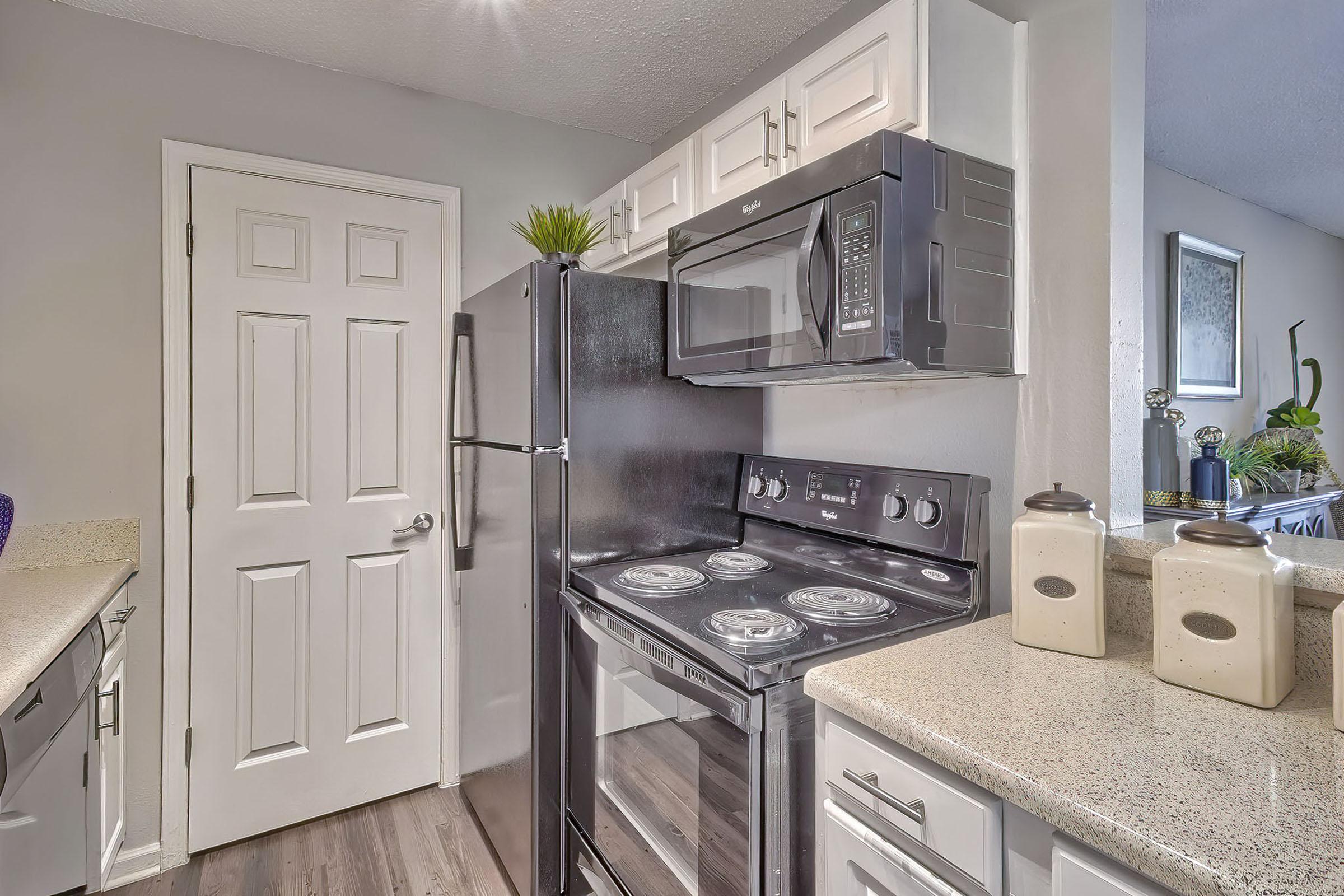 A modern kitchen featuring white cabinetry, a dark black refrigerator, a microwave above a black stove, and a beige countertop. The room has gray walls and a door in the background, with decorative plants and jars on the counter, creating a clean, functional space.