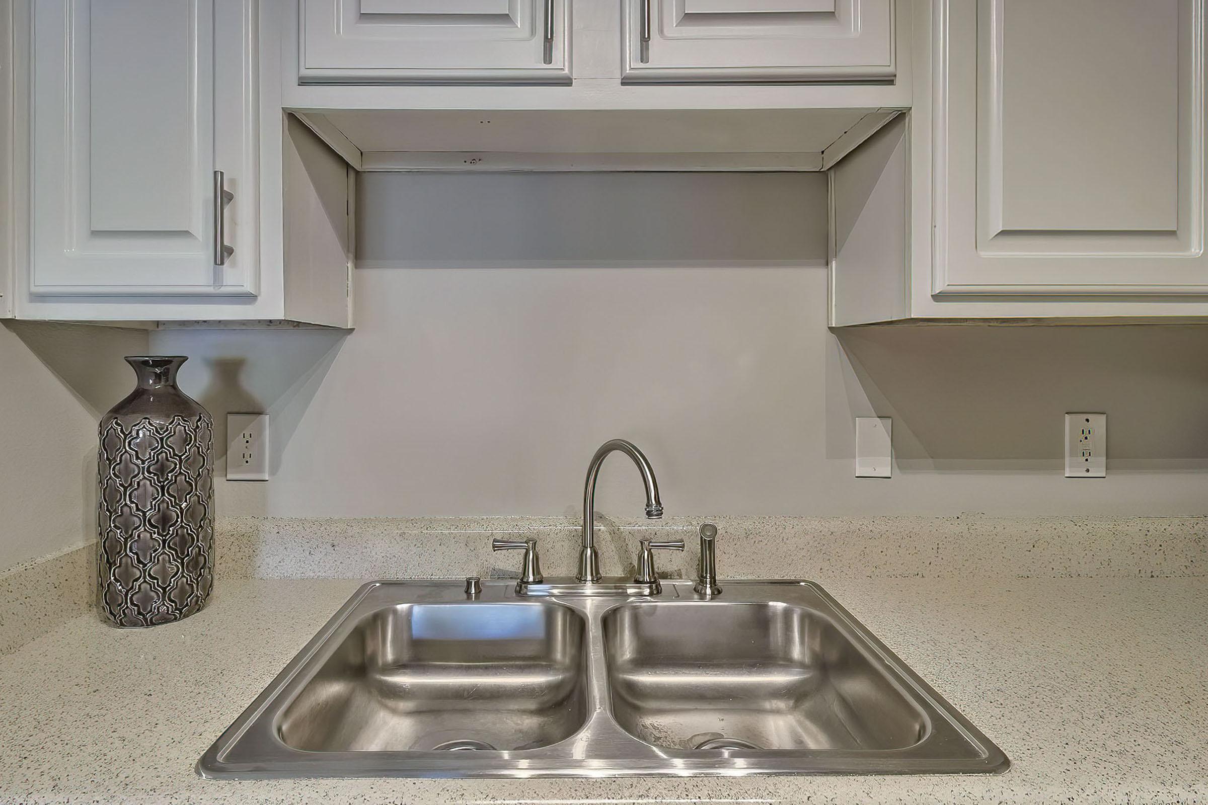 A modern kitchen sink with a double basin, silver faucets, and a decorative gray vase on the left. The countertop is light-colored with speckles, and there are white cabinets above the sink. The wall behind the sink is painted a soft gray, and electrical outlets are visible on either side.