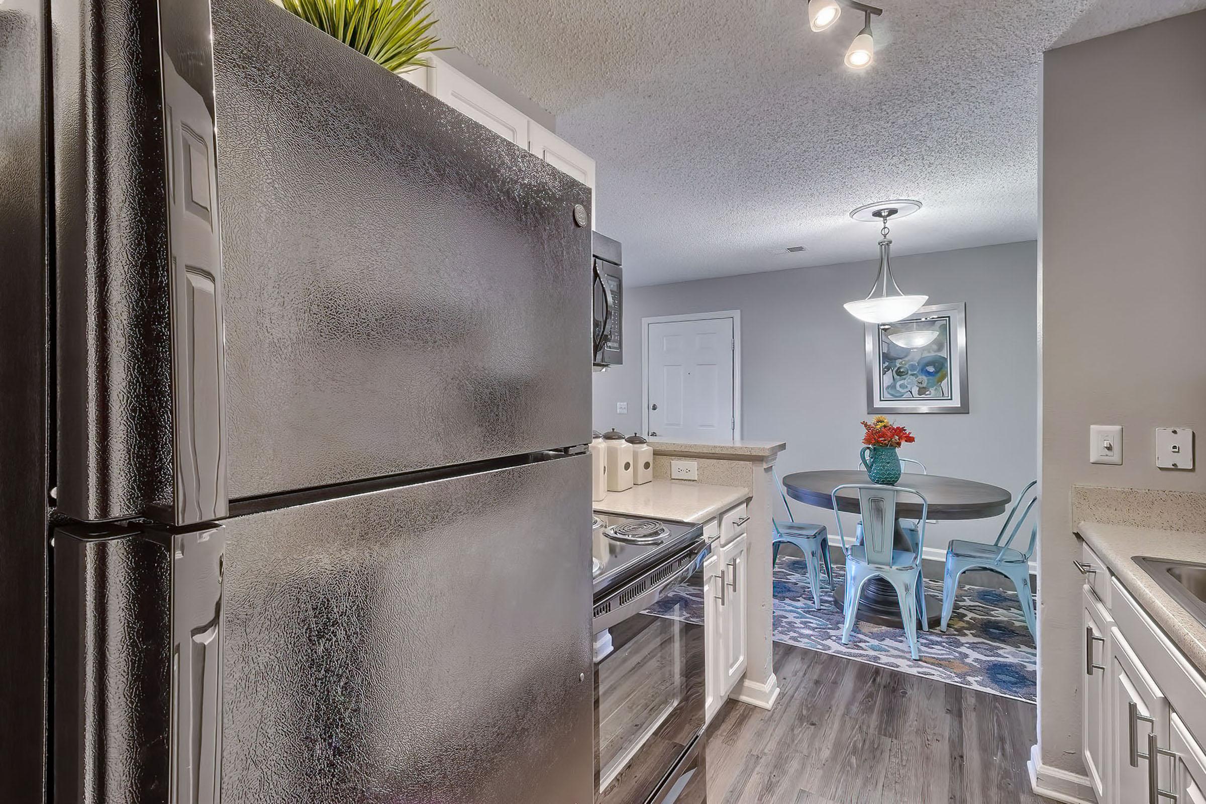 Modern kitchen interior featuring a stainless steel refrigerator, an oven, and a dining area with a round table and blue chairs. The space includes light-colored cabinetry, a potted plant, and decorative items on the countertops. Natural light from a nearby window brightens the setting.