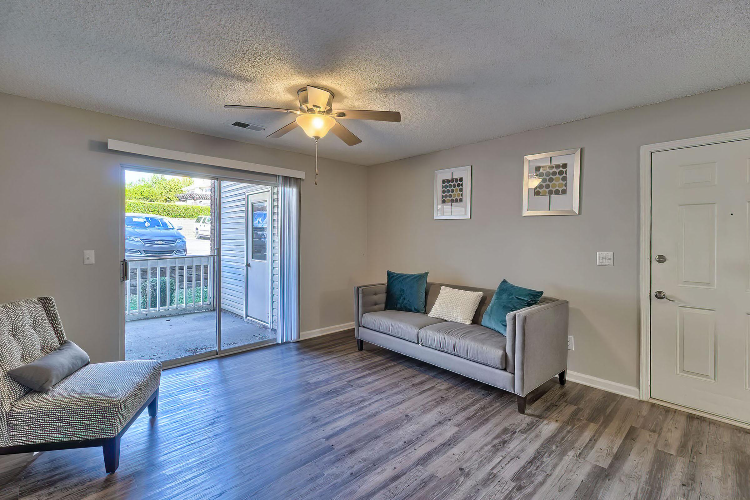 A clean and modern living room featuring a gray sofa with teal cushions, an accent chair, and a ceiling fan. Large sliding glass doors lead to an outside area, while framed artwork decorates the walls. The flooring is a sleek laminate, enhancing the room's contemporary feel.