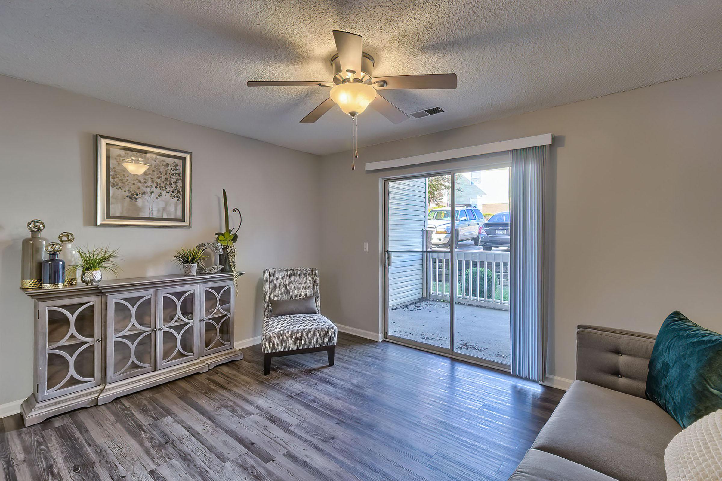 Light-filled living room featuring a gray sofa, a decorative chair, and a stylish gray console table. A ceiling fan adds functionality, and sliding glass doors lead to an outdoor area, enhancing the open feel of the space. Subtle decor elements like plants and wall art create a cozy atmosphere.