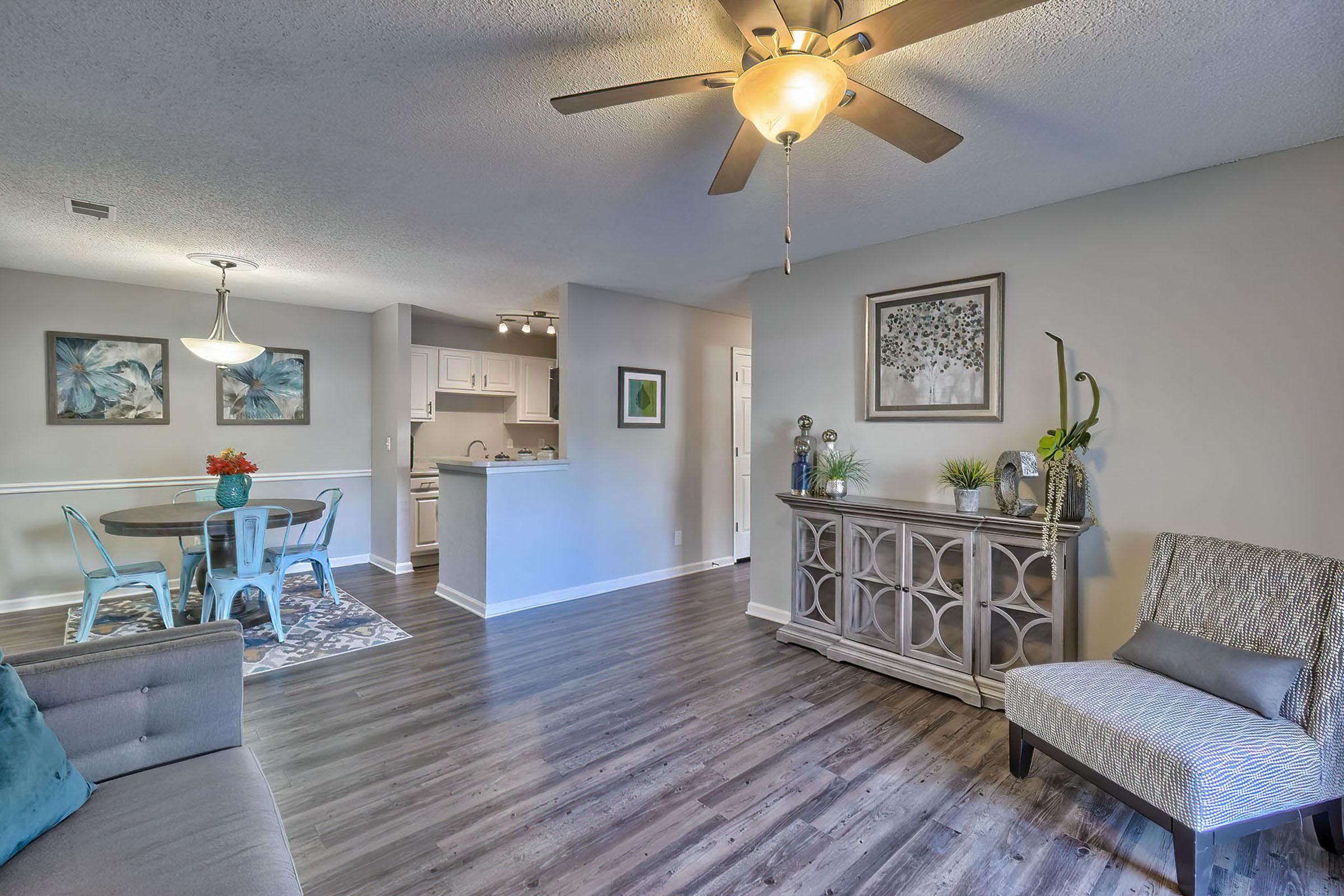 A bright and inviting living space featuring a comfortable couch, a decorative chair, and a stylish sideboard. Light-colored walls and wooden flooring complement the room, while a dining area with a table and chairs is visible in the background, along with a kitchen area. Natural light enhances the cheerful ambiance.