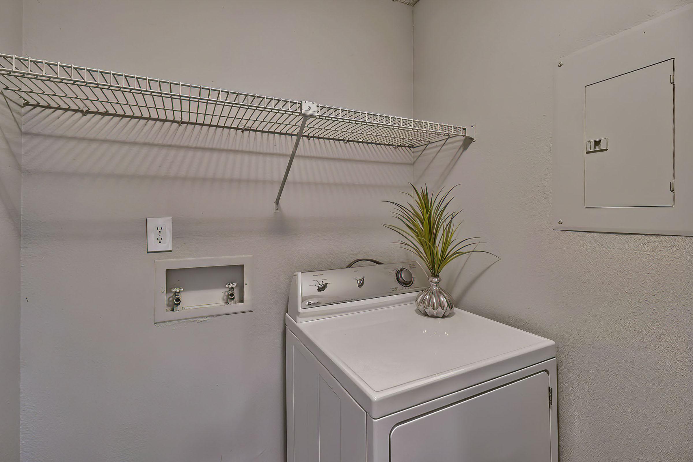 A laundry room featuring a white washing machine on the left, a decorative plant on top, and a wire shelf above. The wall is painted in a light color, and there is an electrical panel on the right. The space is well-lit and tidy, providing a functional area for laundry tasks.