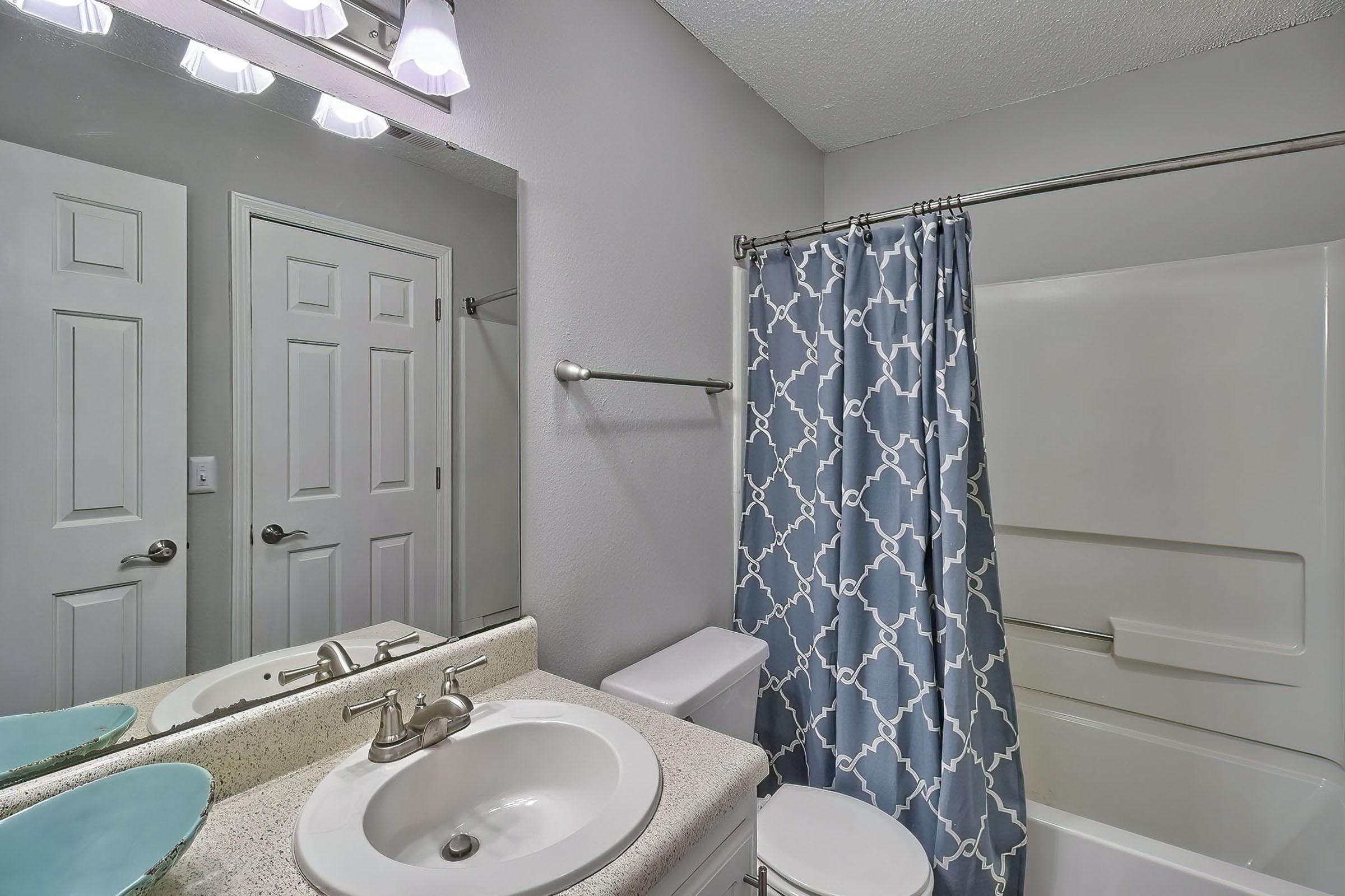 A modern bathroom featuring a white countertop with a sink, a chrome faucet, and a decorative blue shower curtain. A large mirror above the sink reflects the light from four bulbs. There’s a white toilet and a bathtub with a shower fixture, all set against a light gray wall.