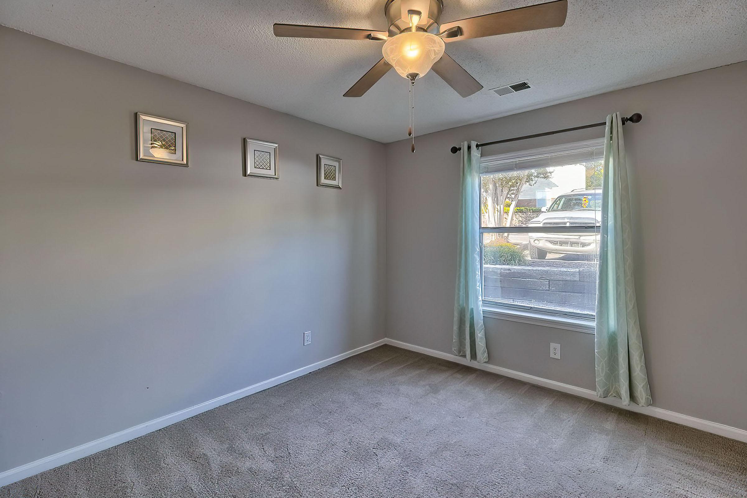 A neutral-colored bedroom with carpet flooring, featuring a ceiling fan, three framed pictures on the wall, and a window with light green curtains. The window shows a view of the outdoors and a glimpse of a vehicle parked outside.