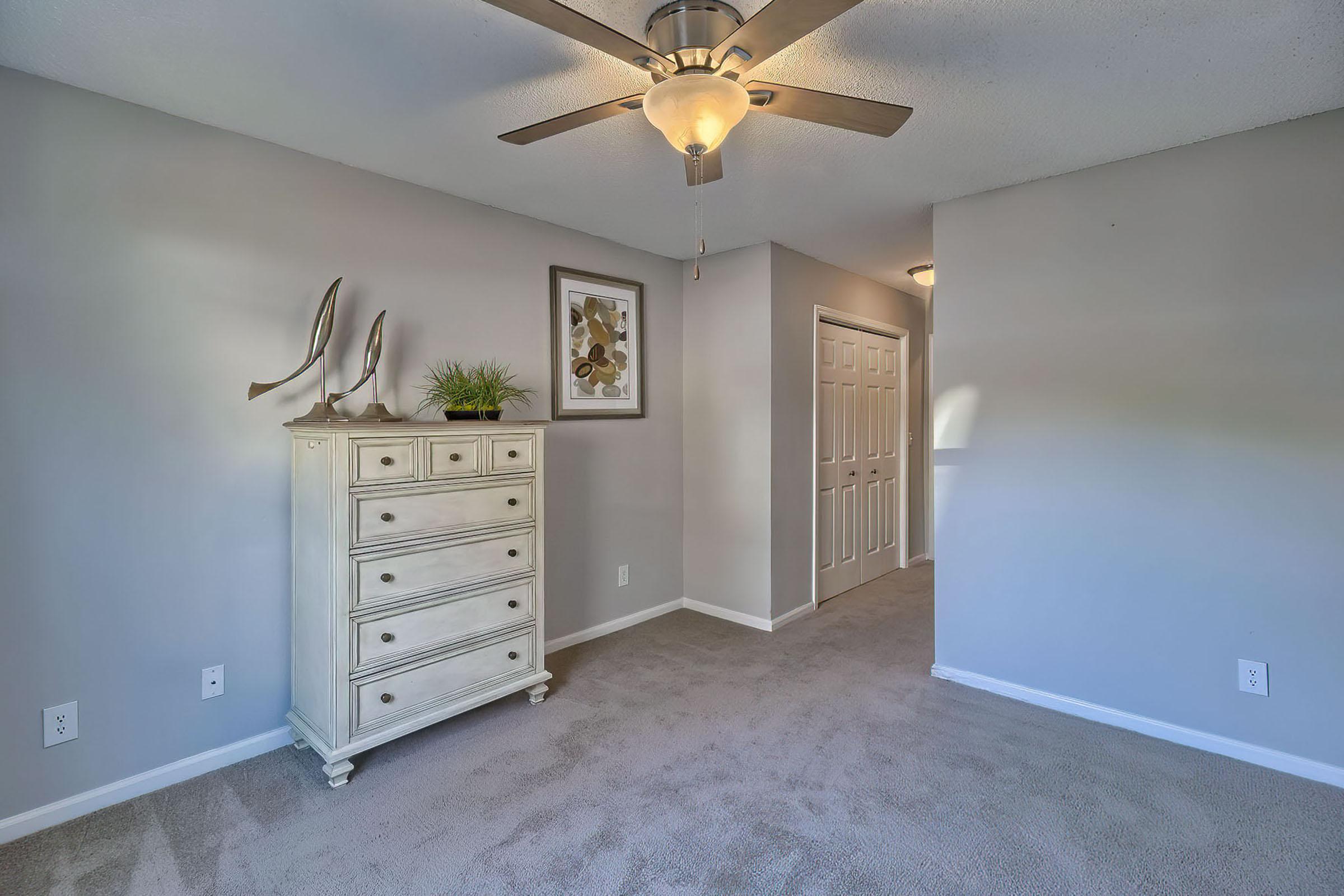 A well-lit bedroom featuring light gray walls and beige carpeting. A decorative white dresser with six drawers stands against one wall, accompanied by a framed piece of art and a small green plant. A ceiling fan is mounted above, and a white door is seen leading to another room in the background.