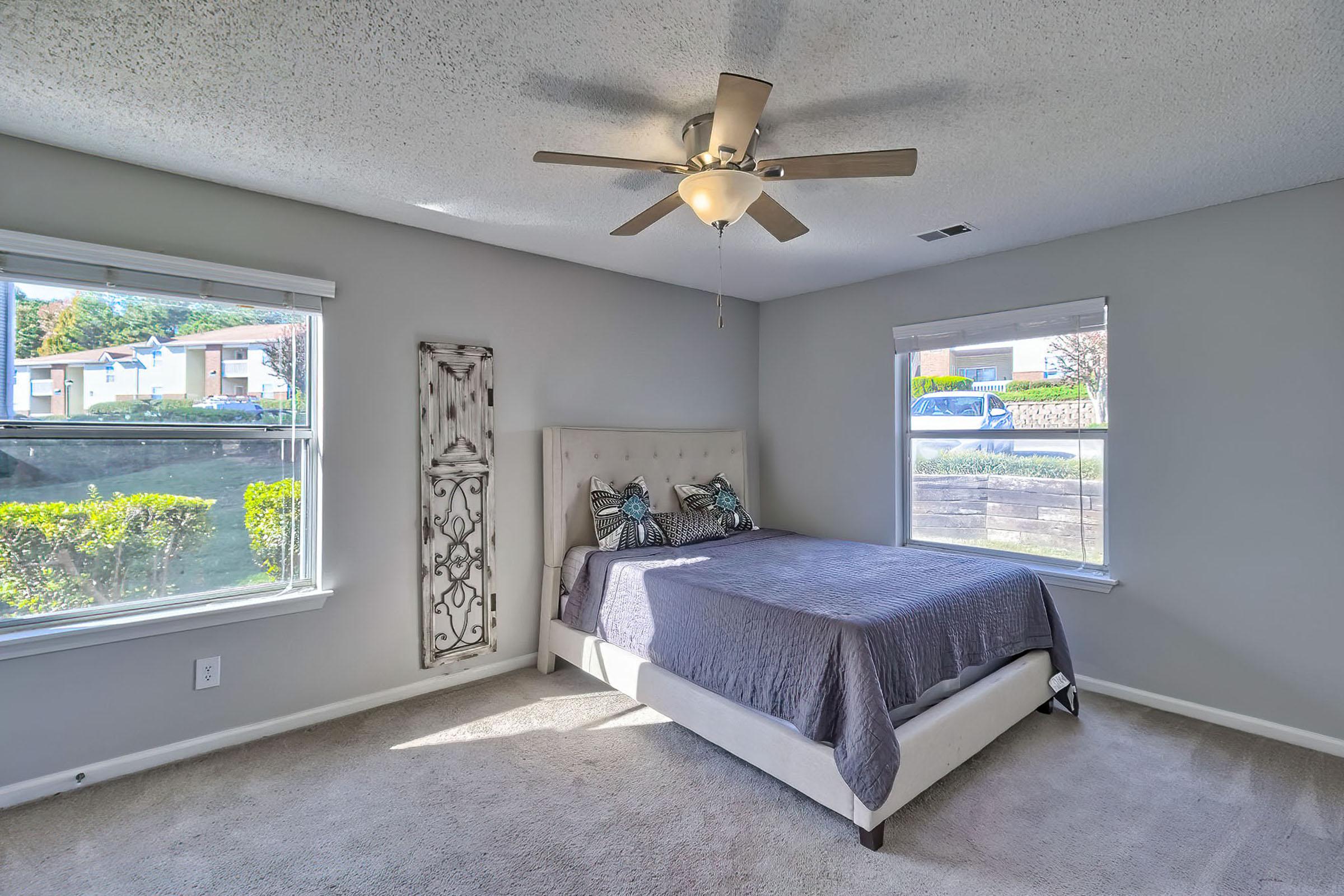 A well-lit bedroom featuring a light gray wall, a plush bed with a decorative headboard, and two windows providing natural light. A ceiling fan is visible above, and there is a decorative wooden panel on the wall. The room has soft carpeting and greenery visible outside the windows.