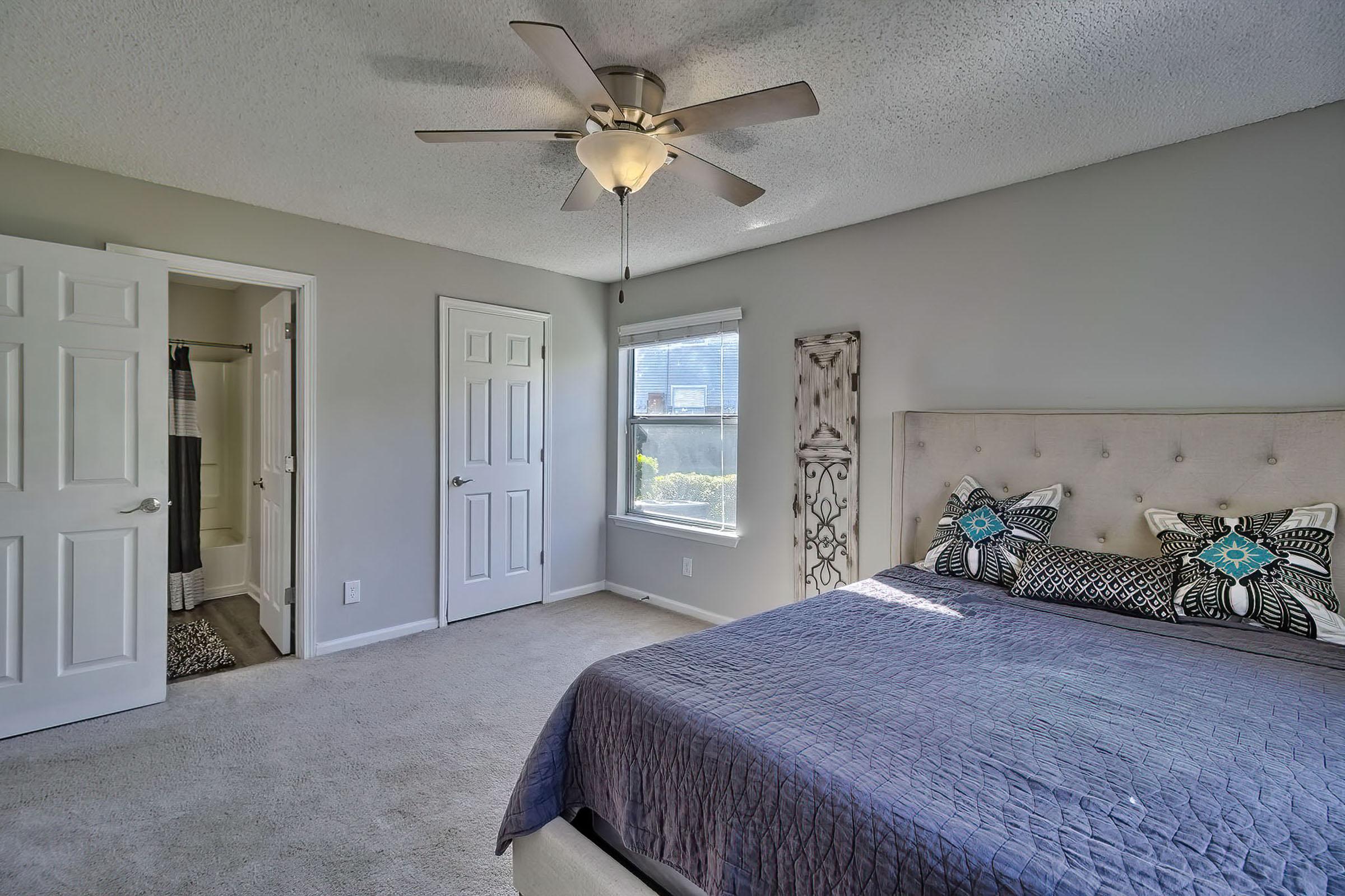 A bright and airy bedroom featuring a neatly made bed with decorative pillows, a ceiling fan, and light-colored walls. There are two closed doors leading to closets, a window with natural light, and an en-suite bathroom visible in the background. The carpeted floor adds a cozy touch.