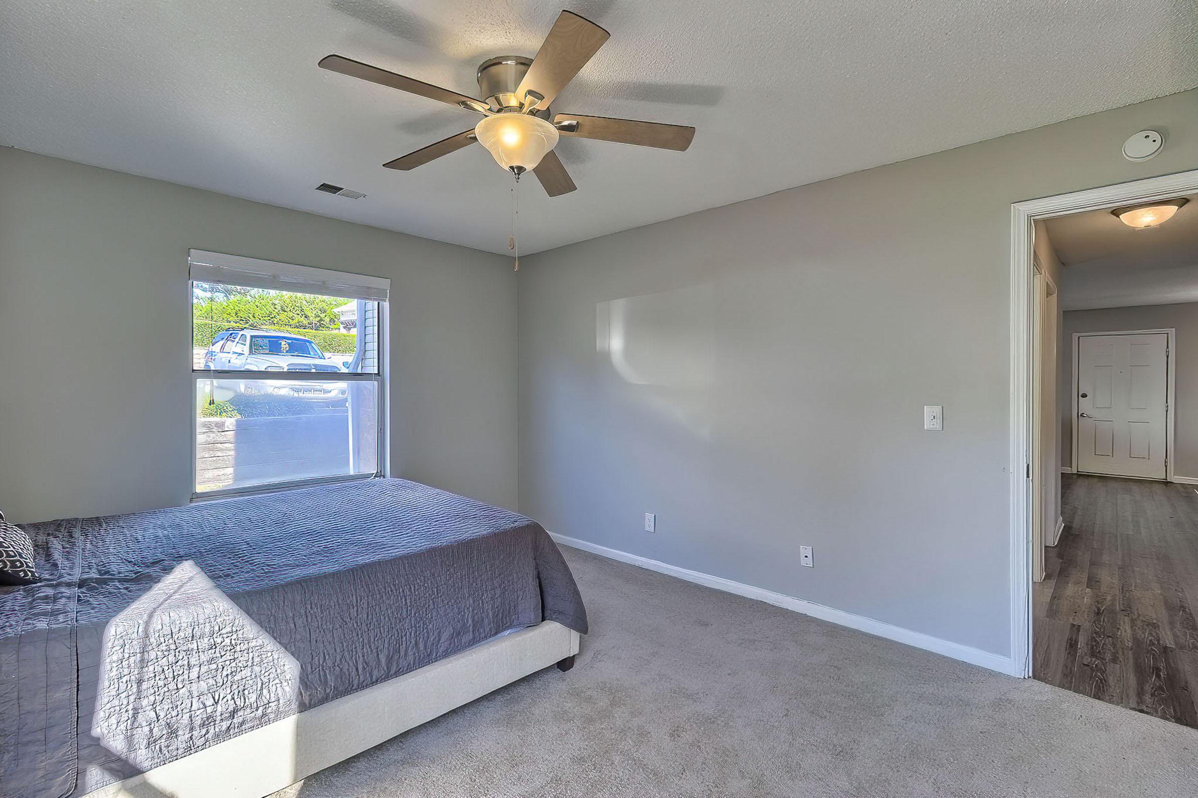 A well-lit bedroom featuring a queen-size bed with a gray quilt, a ceiling fan, and a window showcasing outdoor greenery. The walls are painted light gray, and the room has a carpeted floor. A doorway leads to another room, while a glimpse of a hallway is visible on the right side.