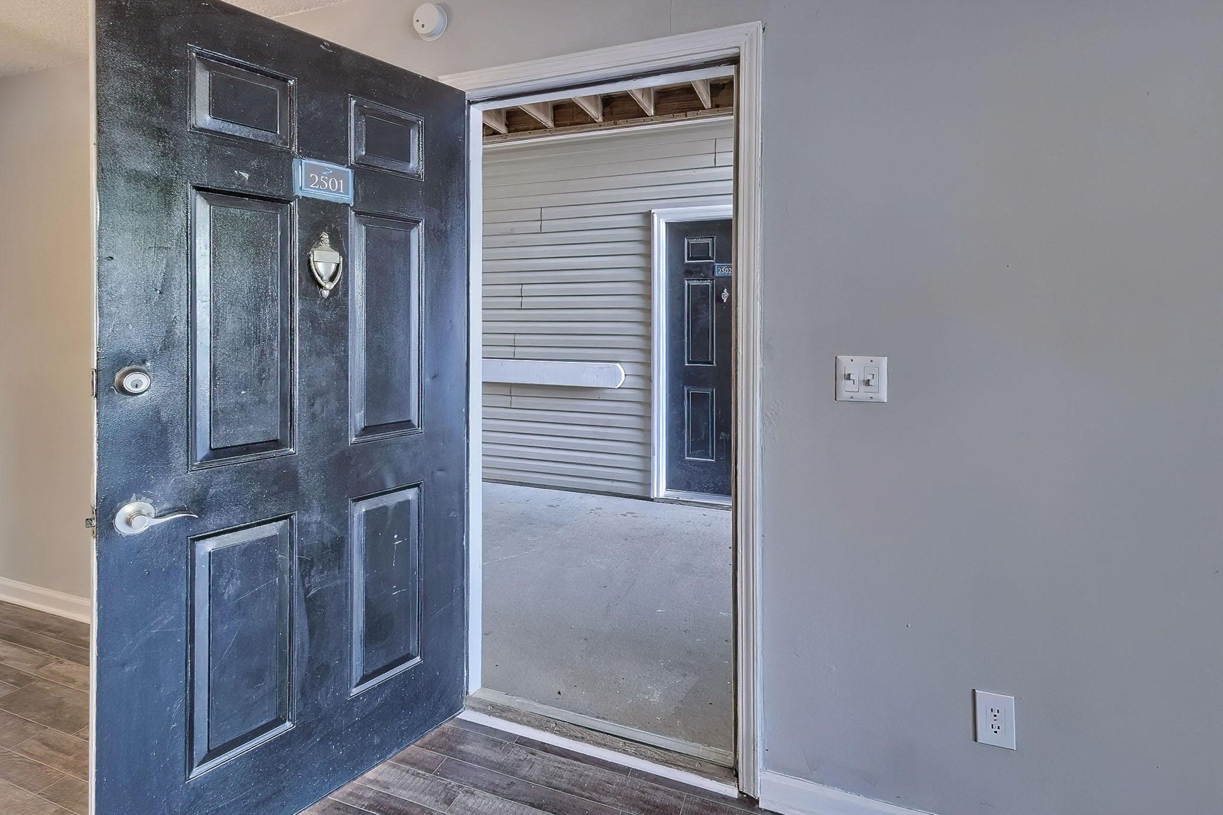 An open black front door leads into a hallway with bare walls and a view of another closed door in the background. The interior has a simple, unfinished look with wooden flooring and exposed ceiling beams. A small wall-mounted light switch is visible near the door frame.