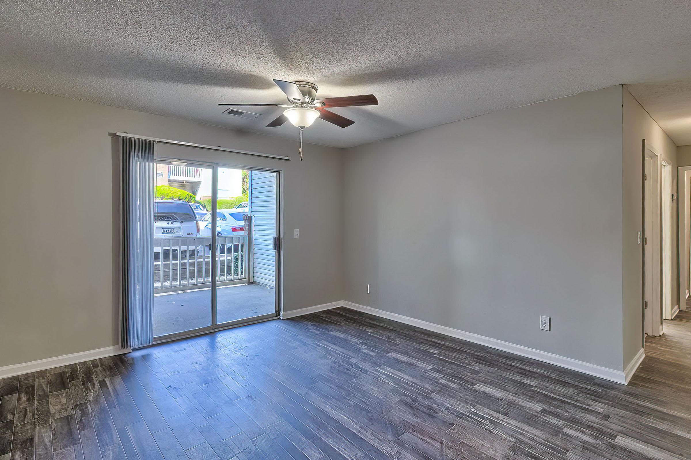 Empty living room with a ceiling fan and sliding glass door leading to a small balcony. Light gray walls and dark wood-like flooring create a modern feel. Natural light enters through the glass door, brightening the space. A doorway on the right leads to another room or hallway.