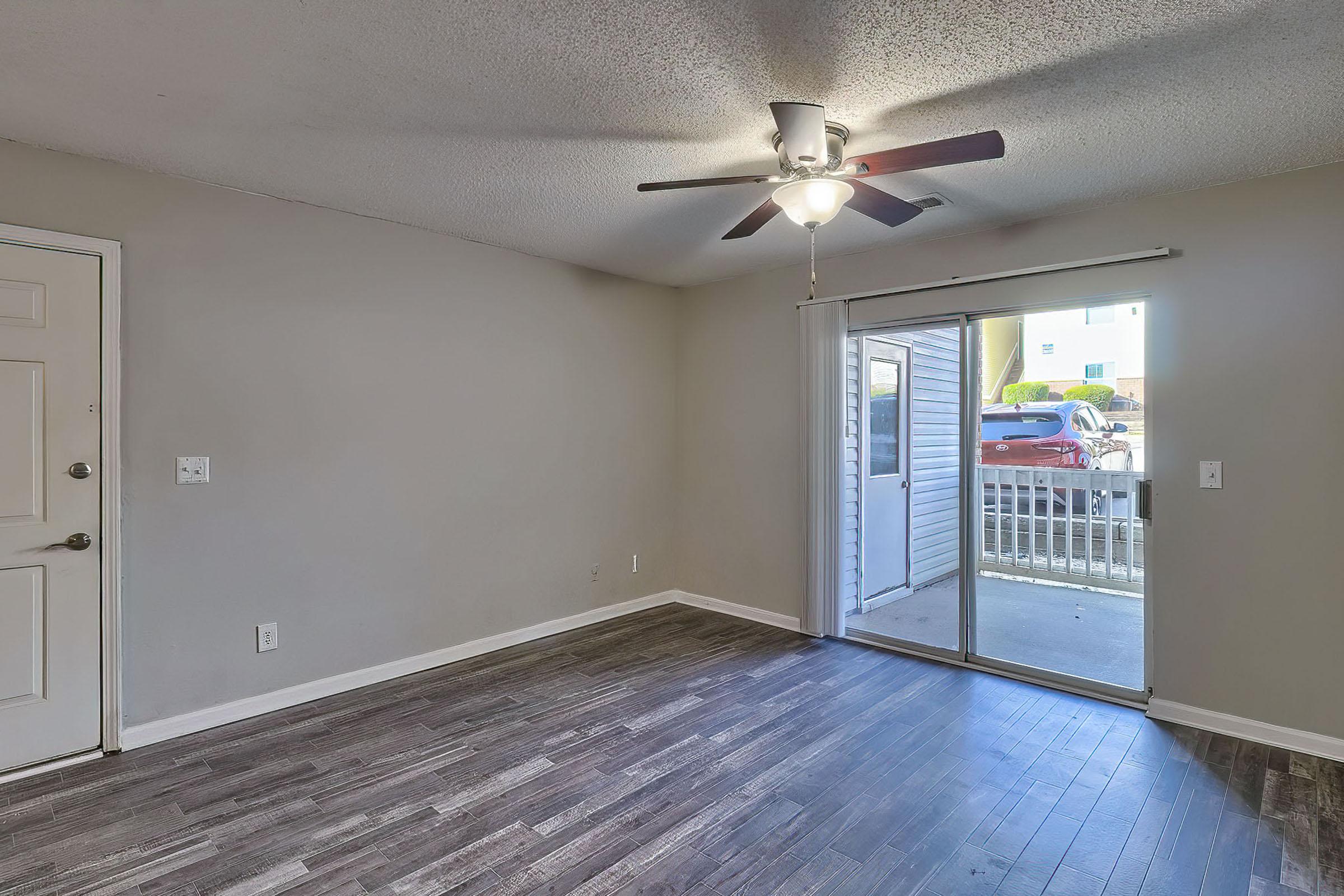 A bright, empty living room featuring a ceiling fan, a sliding glass door leading to an outside area, and dark wood-like flooring. The walls are painted a light gray, creating a spacious and inviting atmosphere. Natural light streams in through the sliding door.